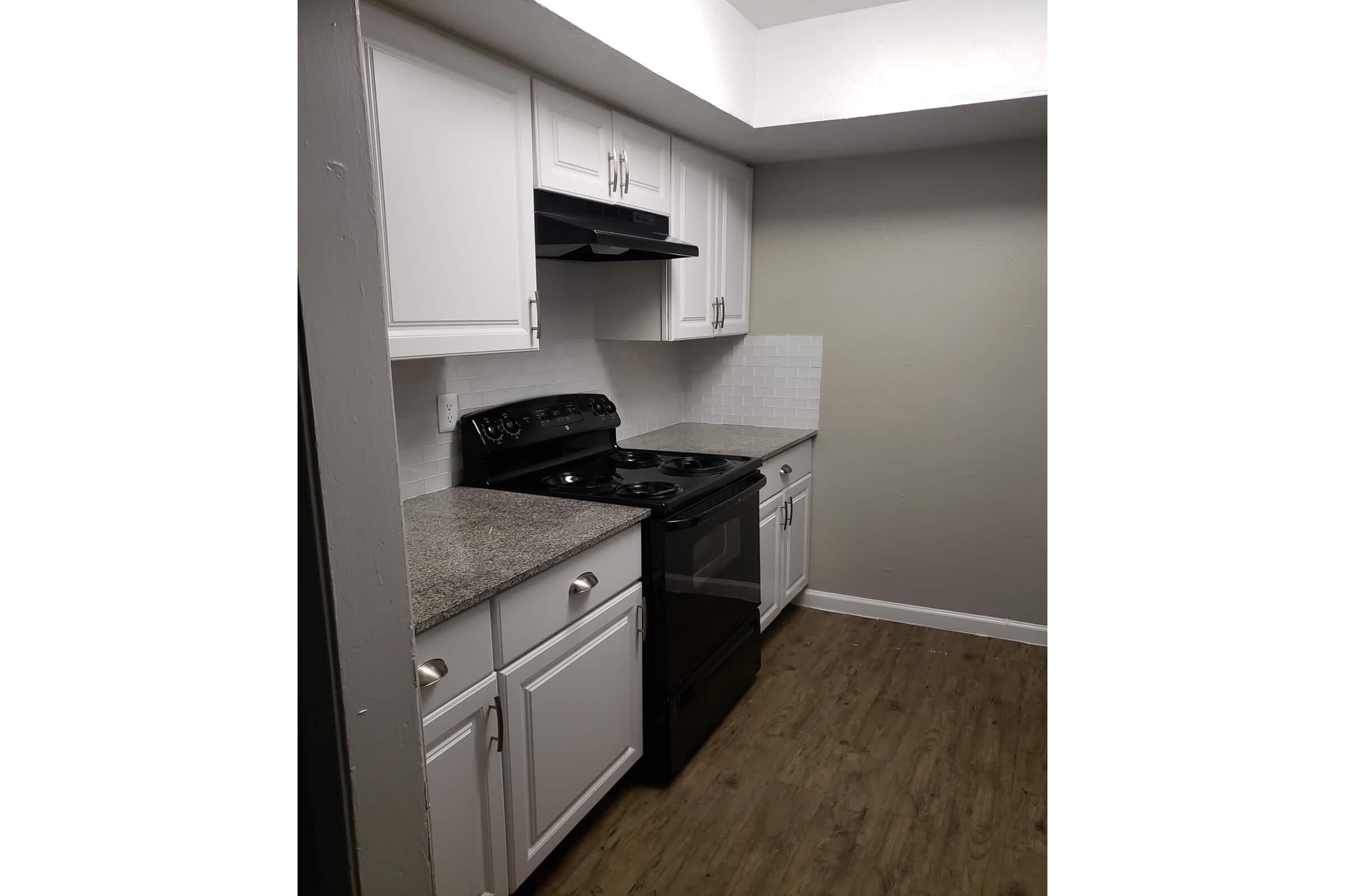 Modern kitchen with white cabinetry, a granite countertop, and black appliances, including a stove and hood. The walls are painted in a light gray, and the flooring appears to be wood-like. Natural light comes from the top, creating a bright and welcoming atmosphere.