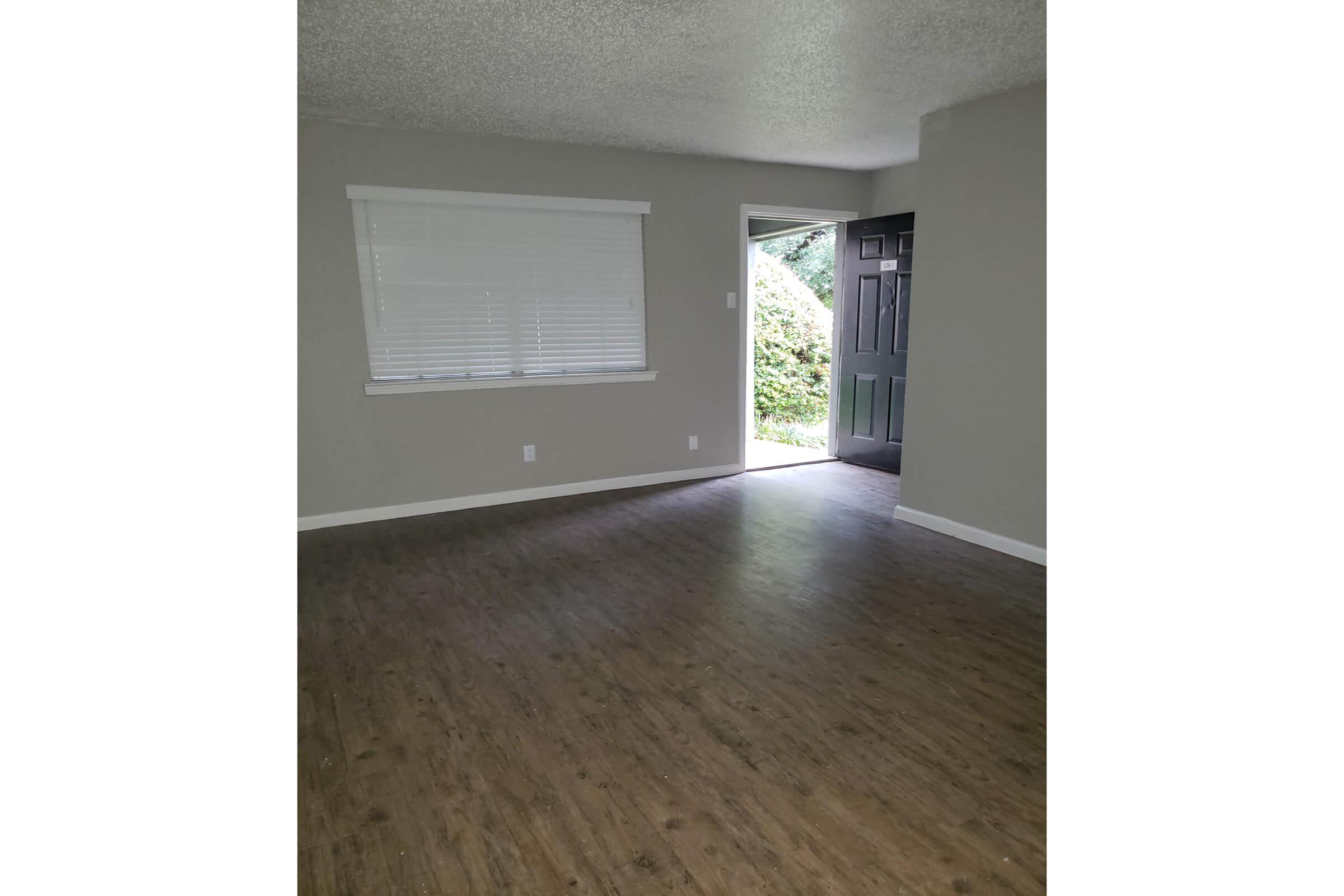 A living room with light gray walls and a wooden floor. A window with white blinds is situated on the left, while an open front door reveals greenery outside. The space is empty and well-lit, creating a welcoming atmosphere.