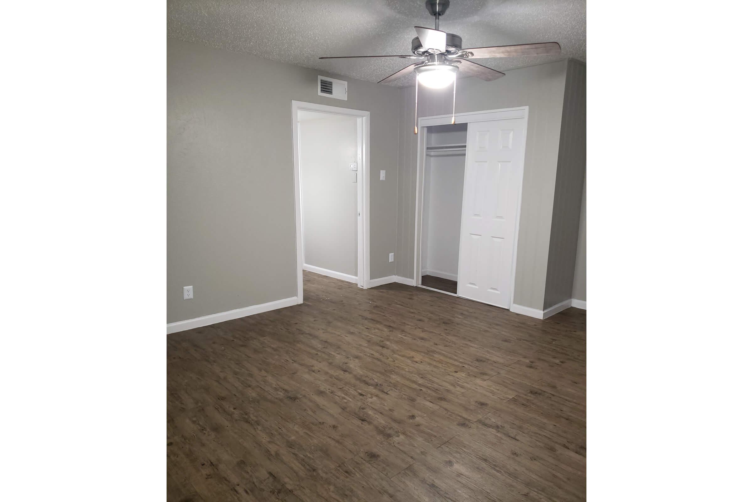 A spacious, empty room with light gray walls and a ceiling fan. The floor features a wood-like laminate. Two doorways lead to adjacent areas, one with a closet. Natural light brightens the space, creating a clean and inviting atmosphere, ideal for various interior designs.