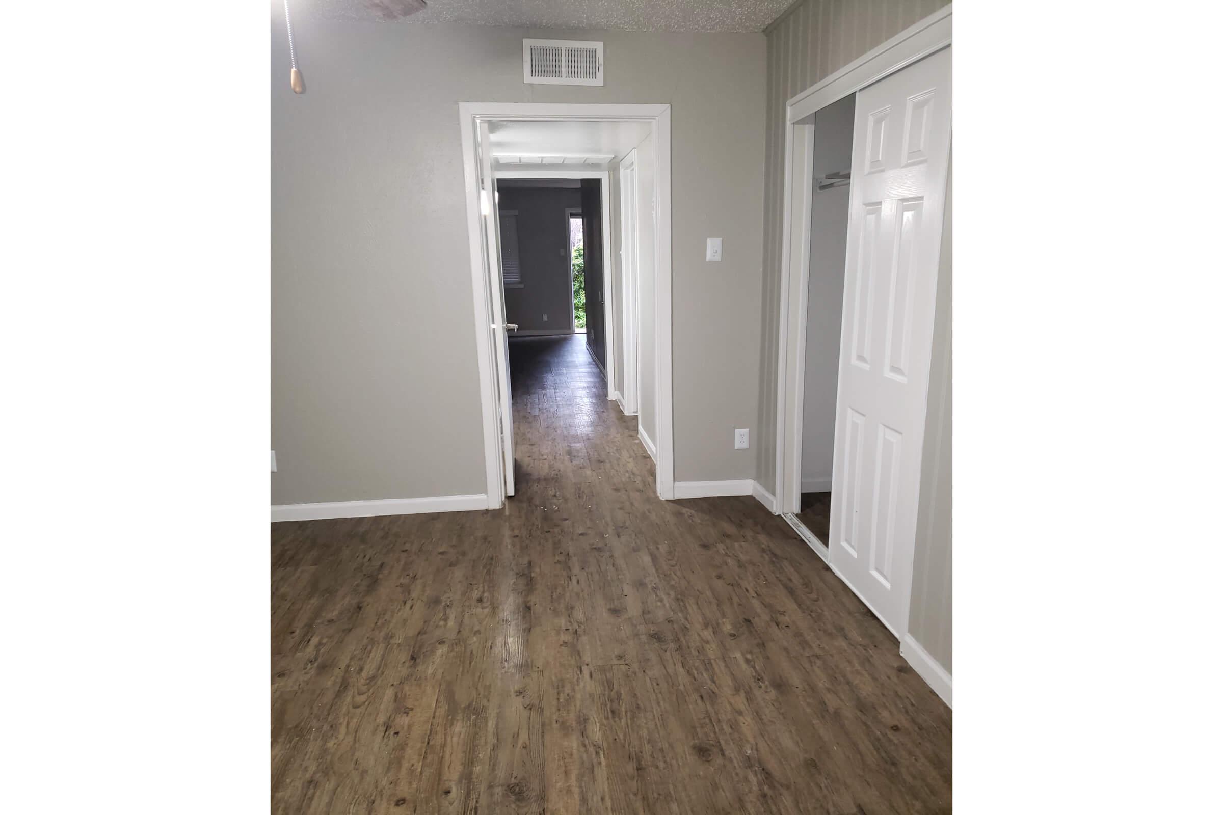 A view of a hallway featuring light-colored walls and a wooden floor, with doors on either side leading to rooms. Visible are two open doors, one on the left and one on the right, and a glimpse of a hallway extending further back. Natural light is coming from the end of the hallway.