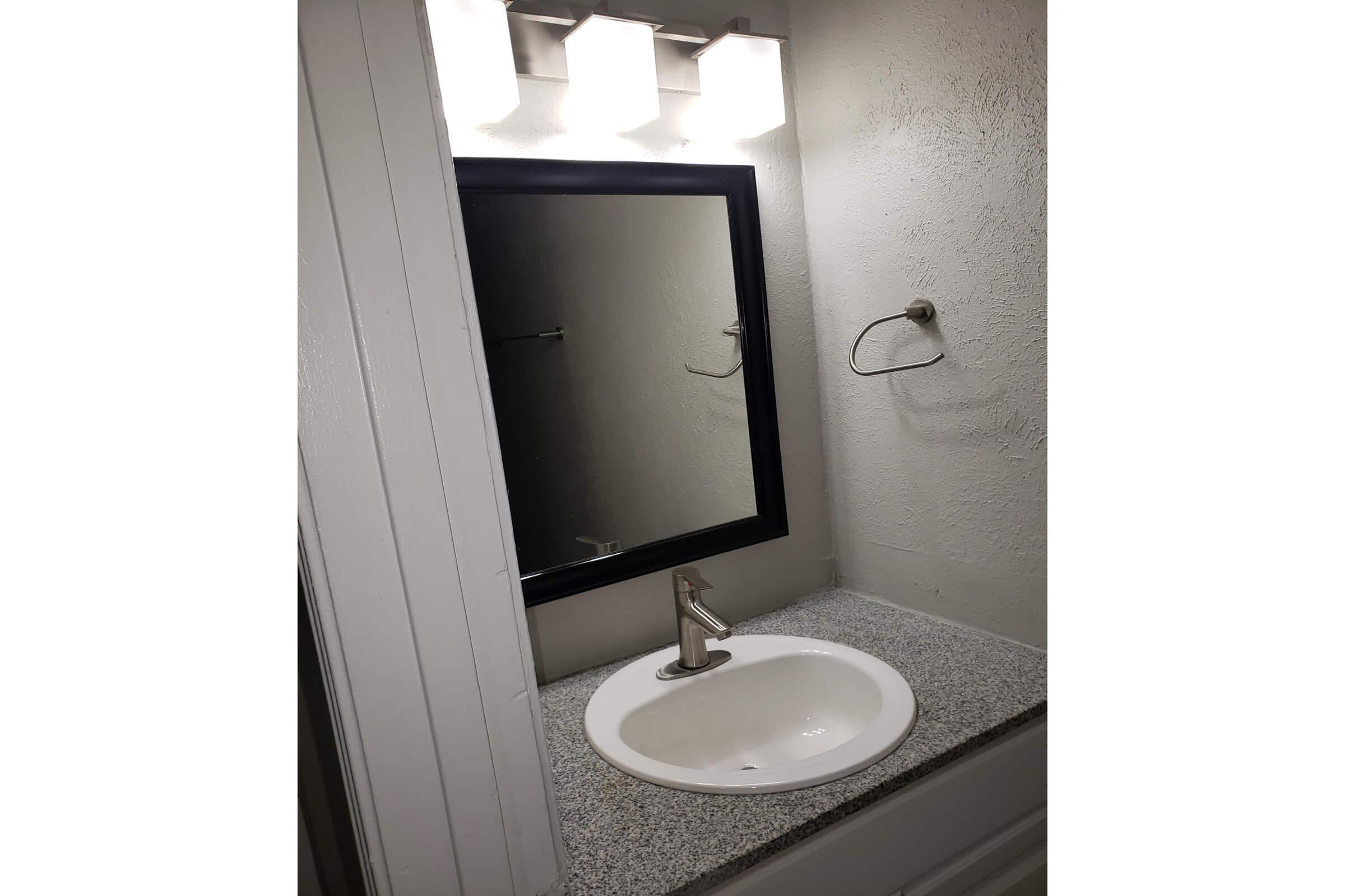 Bathroom sink area featuring a white oval basin, a modern chrome faucet, and a light-colored granite countertop. A rectangular mirror with a black frame hangs above the sink. Three light fixtures are mounted above the mirror, and a towel holder is attached to the wall on the right.