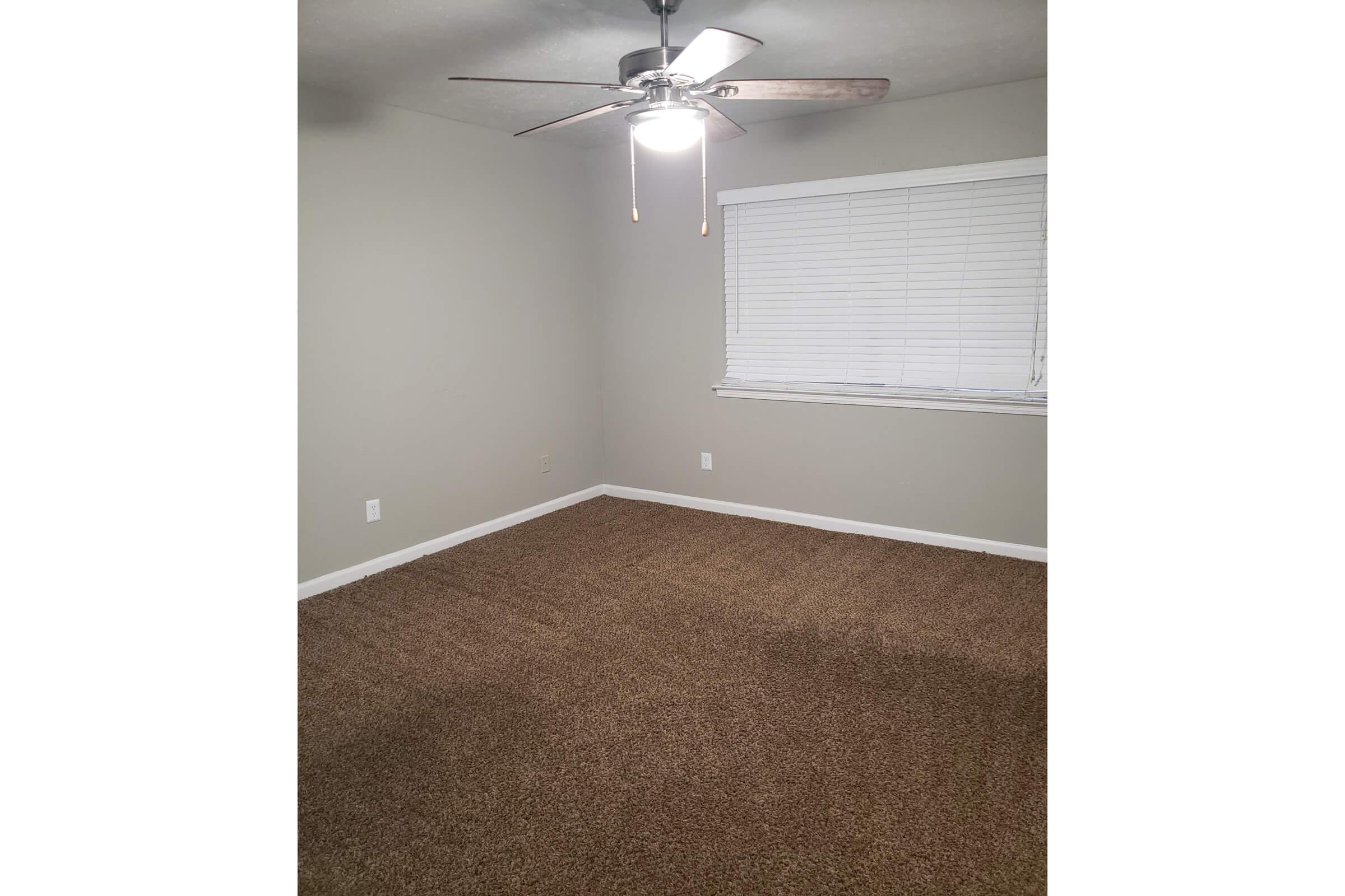 A sparsely furnished room featuring beige carpet, light gray walls, and a ceiling fan with a light fixture. The window has white blinds, and the overall space is bright and uncluttered, creating a simple and clean aesthetic.