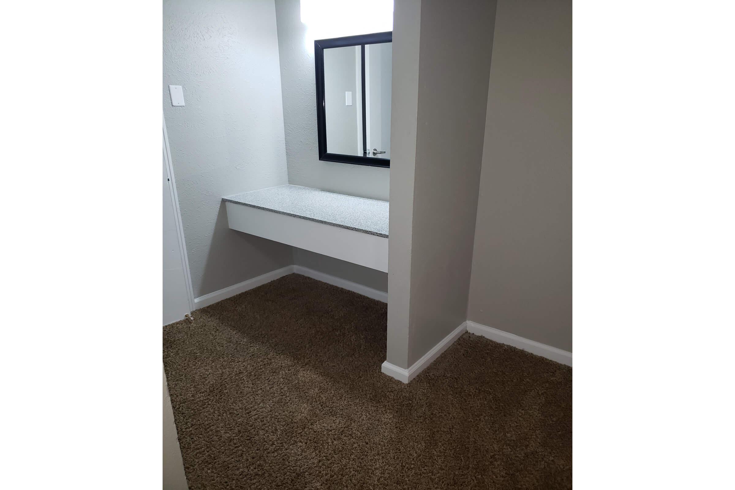 A small, well-lit room with gray walls and a carpeted floor. There is a rectangular vanity with a smooth countertop and a black-framed mirror above it. The space is simple and uncluttered, with a door visible on the left side.