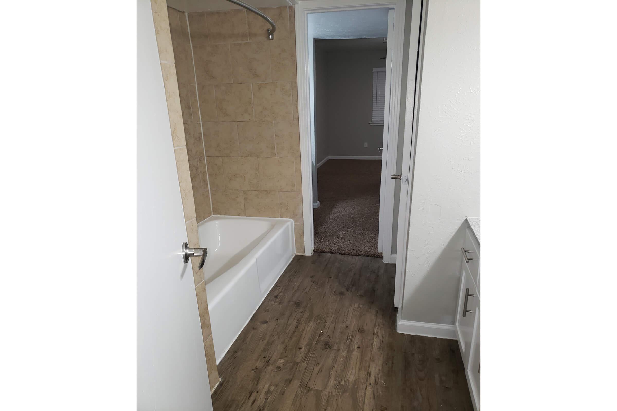 A view of a bathroom featuring a bathtub with a showerhead, light-colored tiled walls, and a wooden floor. The bathroom connects to a carpeted room visible through an open door. White cabinets are present along one wall, and the space is well-lit with a neutral color palette.