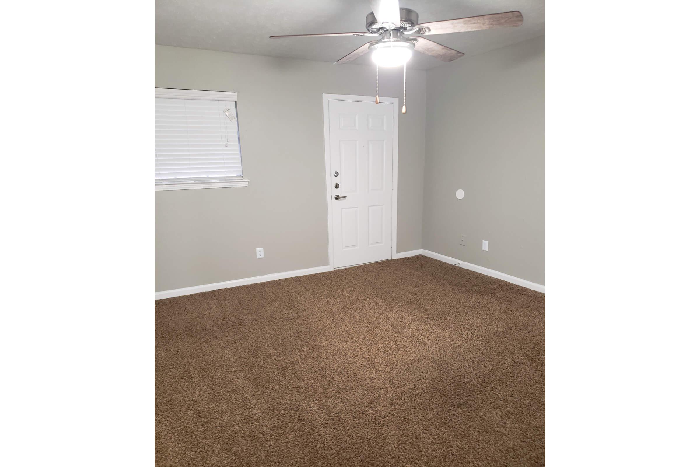 A small, empty room with light gray walls and a ceiling fan. The floor is covered in beige carpet. A window with white blinds is on the left wall, and a white door is centered on the right wall. The room has a simple, uncluttered appearance, making it suitable for various purposes.