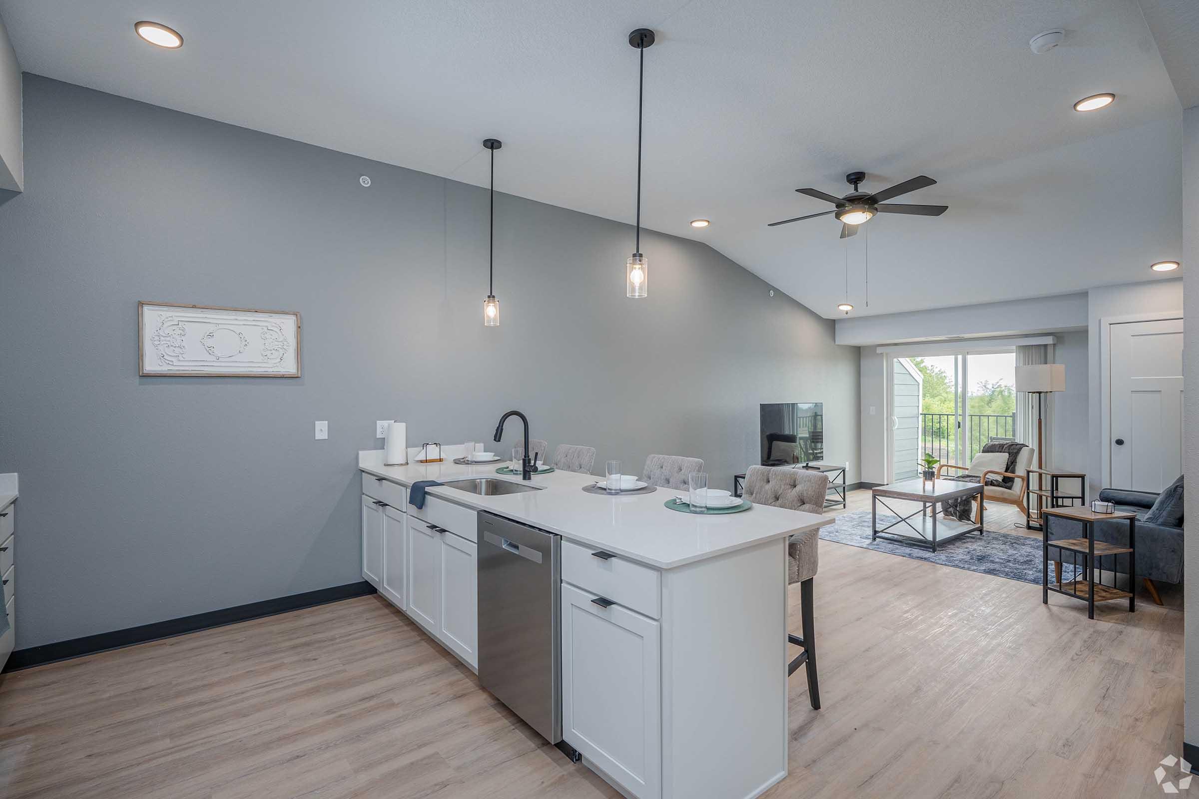 Modern kitchen with an island featuring a sink and dishwasher, surrounded by white cabinetry. The open layout leads to a cozy living area with a large window, comfortable seating, and a decorative rug. Neutral color palette and stylish fixtures enhance the inviting atmosphere.