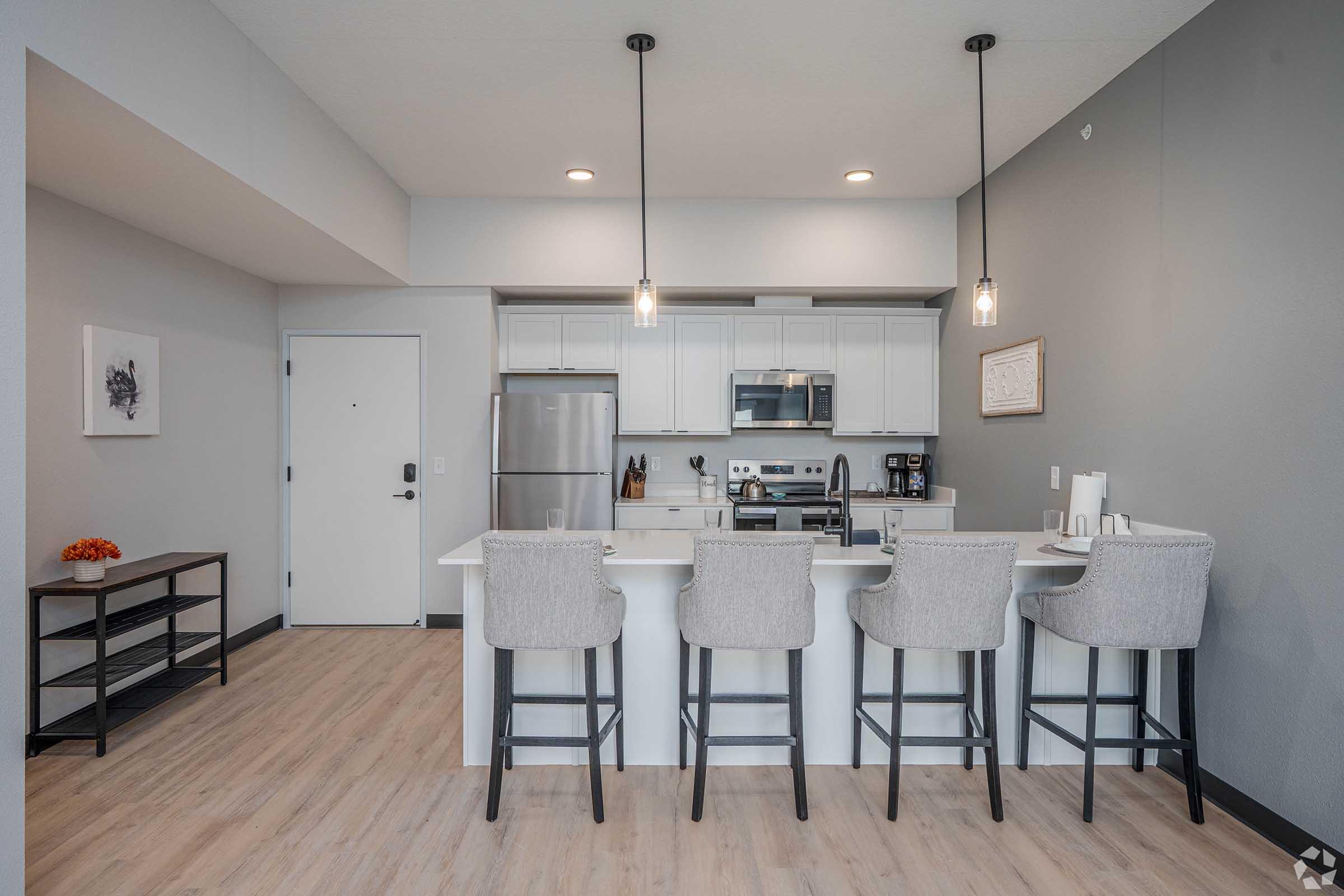 Modern kitchen in an open-concept space featuring a large island with four high-backed stools, stainless steel appliances, white cabinetry, and light wood flooring. Natural light illuminates the area, enhancing the contemporary decor. A minimalist entryway is visible in the background.