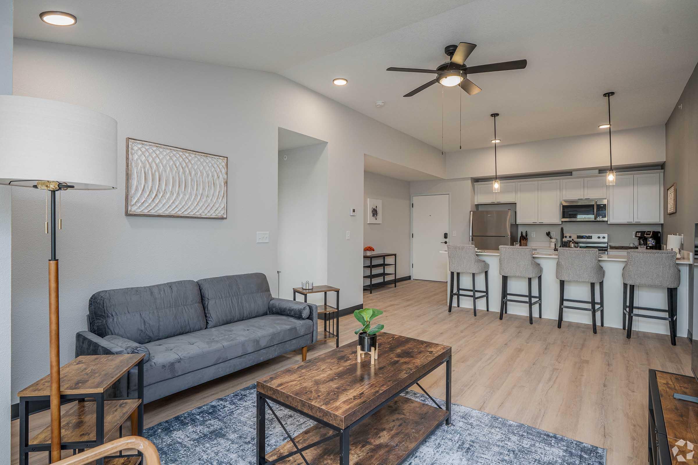 A modern, bright living space featuring a gray sofa, wooden coffee table, and a lamp. The open kitchen is visible in the background with bar stools and stainless-steel appliances. Light hardwood floors and neutral walls create a contemporary atmosphere, complemented by decorative artwork.