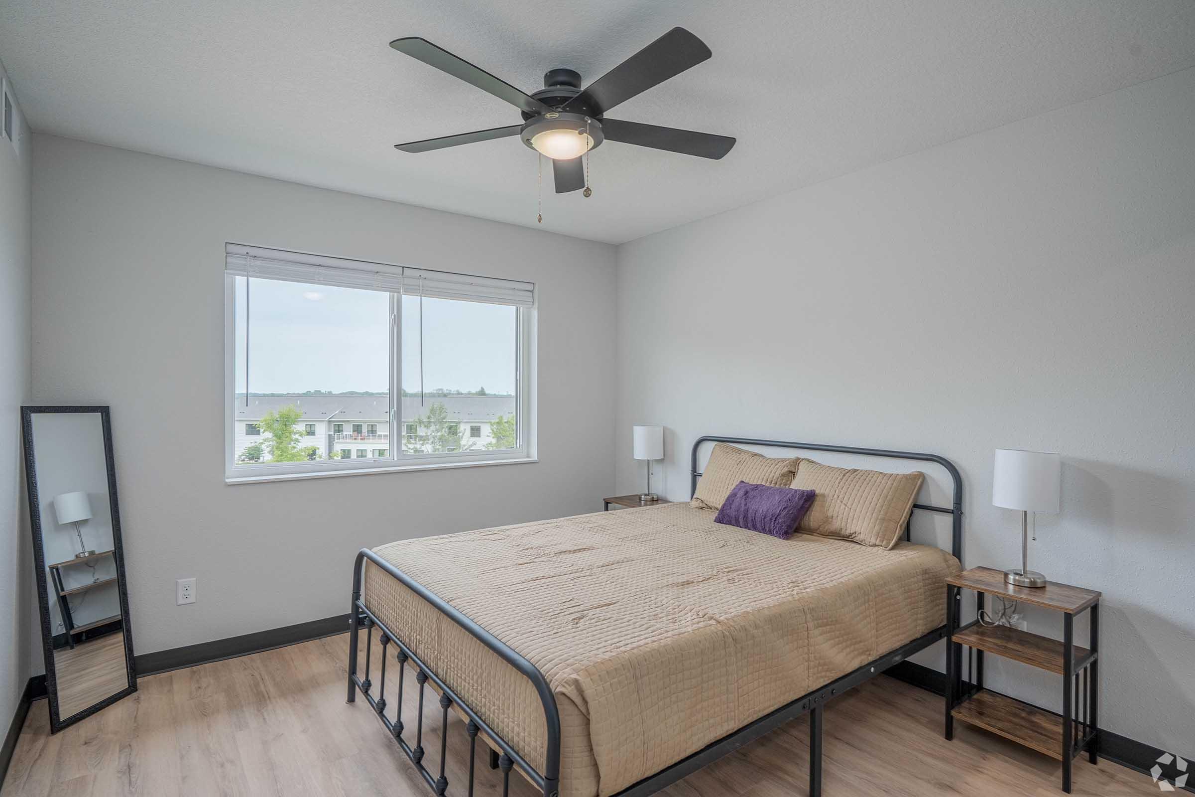 A modern bedroom featuring a metal bed frame with a beige comforter, a bedside table with a lamp on each side, a full-length mirror, and a large window that offers a view of the outdoors. The walls are painted light gray, and the flooring is wooden. A ceiling fan is installed above.