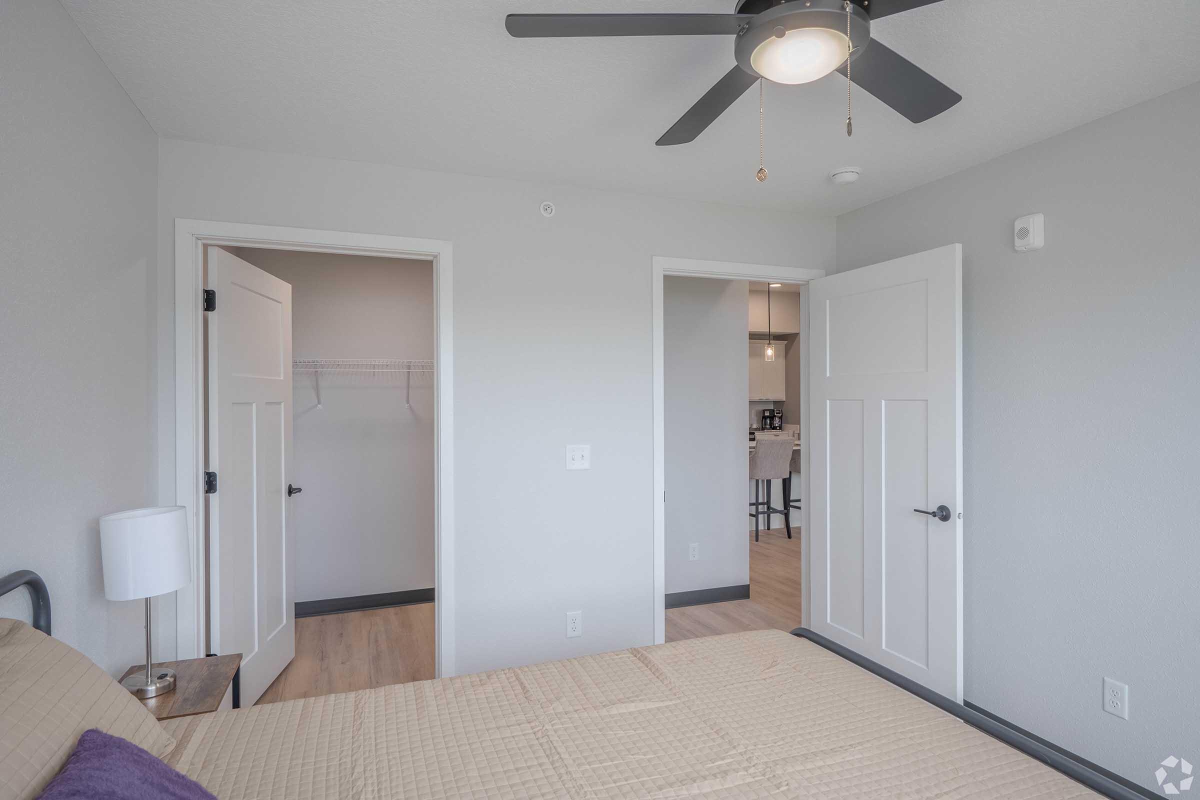 A well-lit bedroom featuring a bed with a beige bedspread, a ceiling fan, and two doors on opposite walls leading to what appears to be a closet and another room. The walls are painted light gray, and the flooring is a light wood finish, creating a modern and clean look.