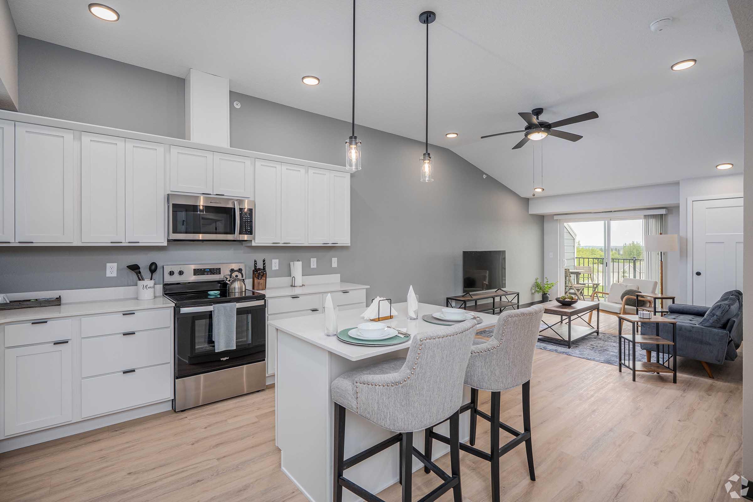 Modern kitchen with white cabinetry, stainless steel appliances, and a large island with seating. The open layout connects to a cozy living area featuring a blue sofa, a TV, and a balcony with outdoor views. Bright lighting complements the contemporary design and neutral color palette.