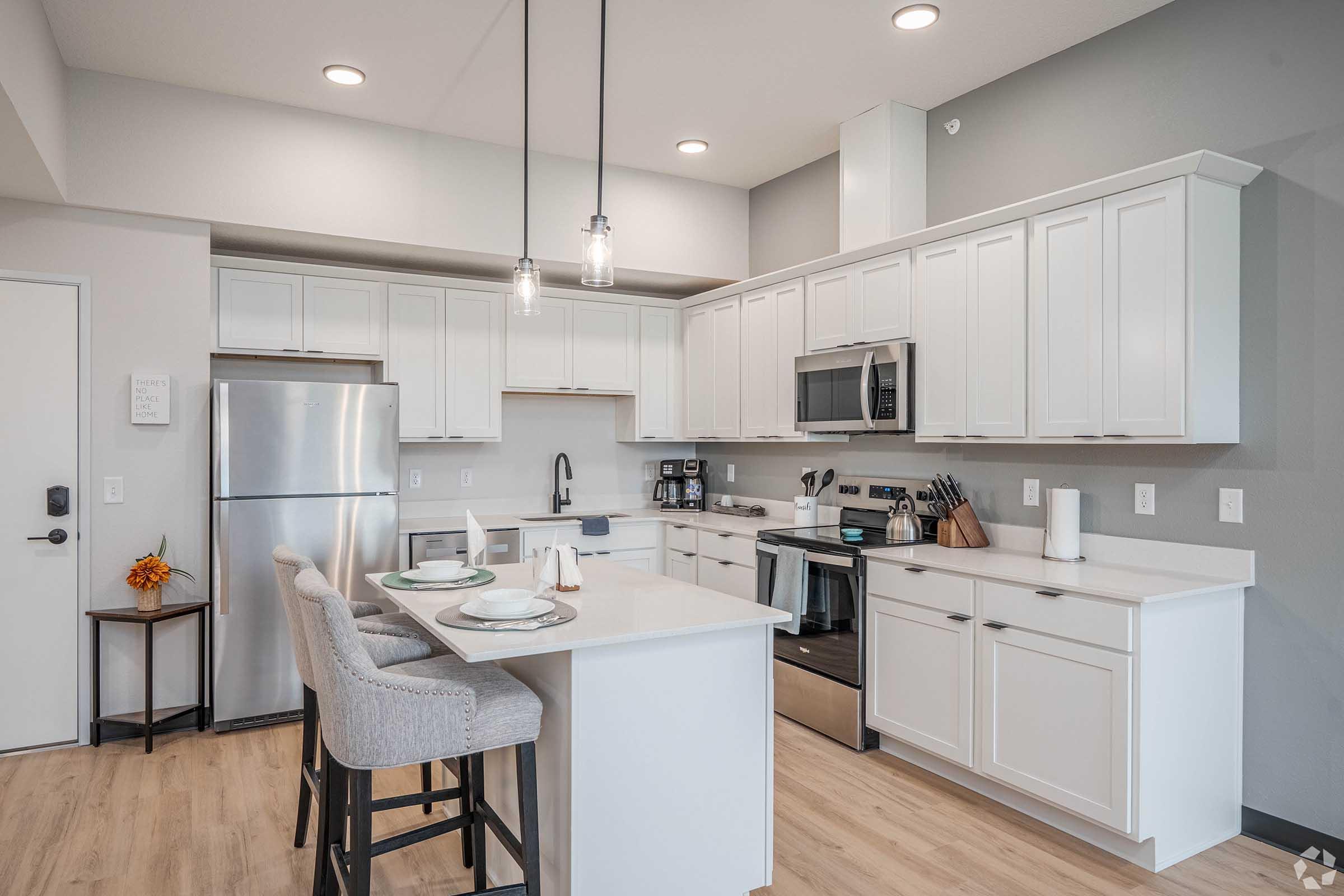 A modern kitchen featuring white cabinetry, stainless steel appliances, a center island with bar stools, and neutral-toned decor. Pendant lights hang above the island, and there is a window allowing natural light. The overall ambiance is clean and contemporary.