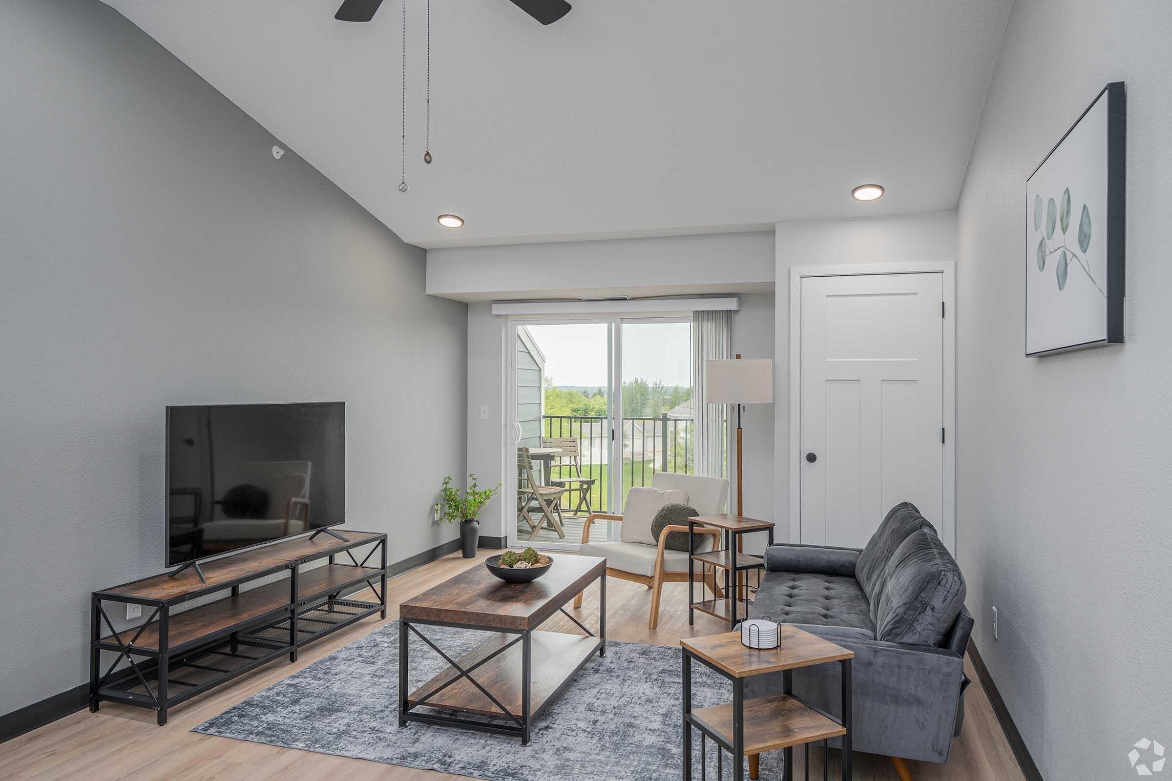 Modern living room featuring a gray color scheme, a comfortable gray sofa, a sleek coffee table, a media console with a television, and a floor lamp. A sliding glass door opens to a balcony with outdoor seating, allowing natural light to brighten the space. Decor includes minimalist artwork and indoor plants.