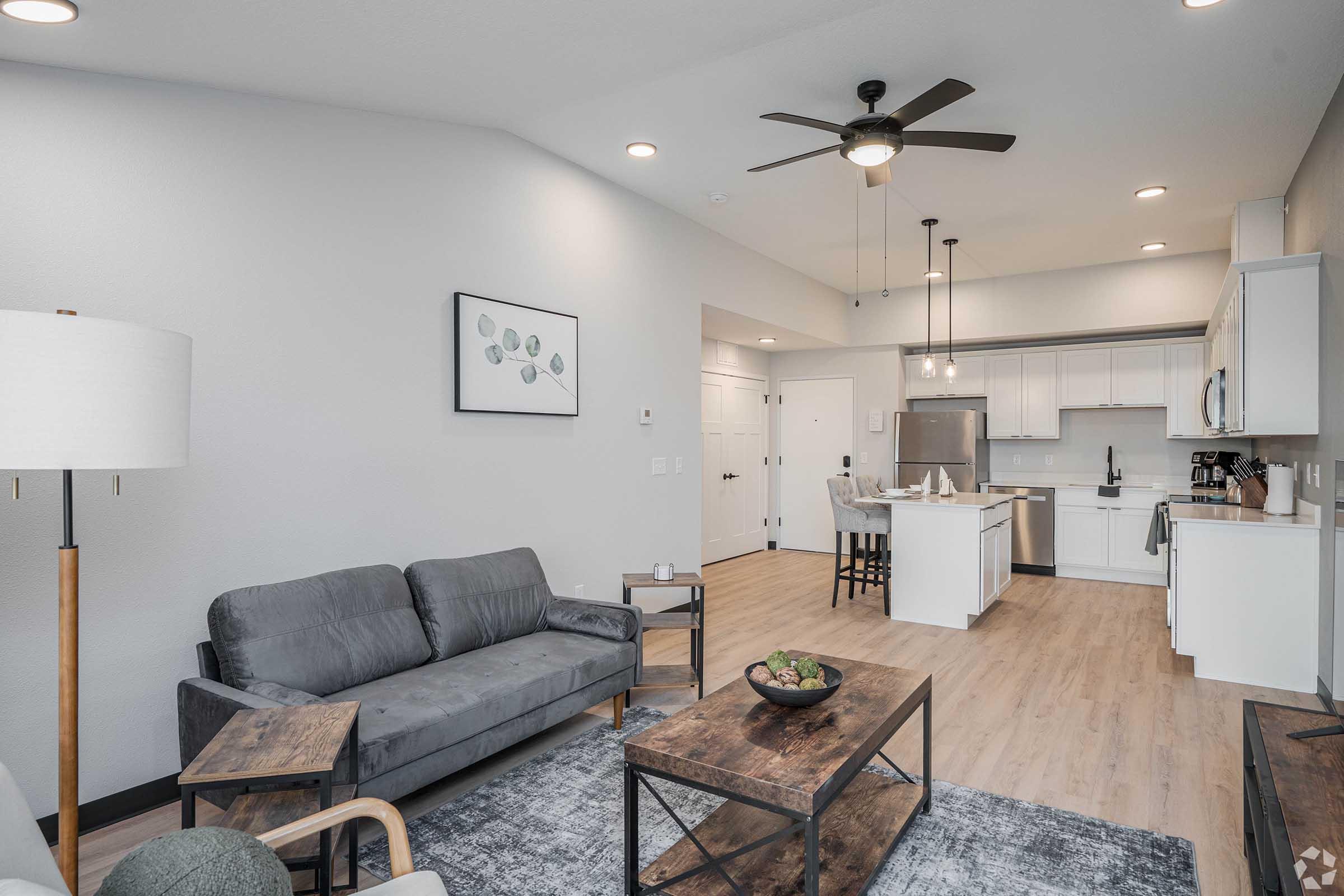 A modern living room featuring a gray sofa and two side tables, a decorative coffee table, and a light fixture. The space has a ceiling fan and is adorned with a wall art piece. In the background, a kitchen area with white cabinets and bar stools is visible, highlighting a clean and contemporary design.