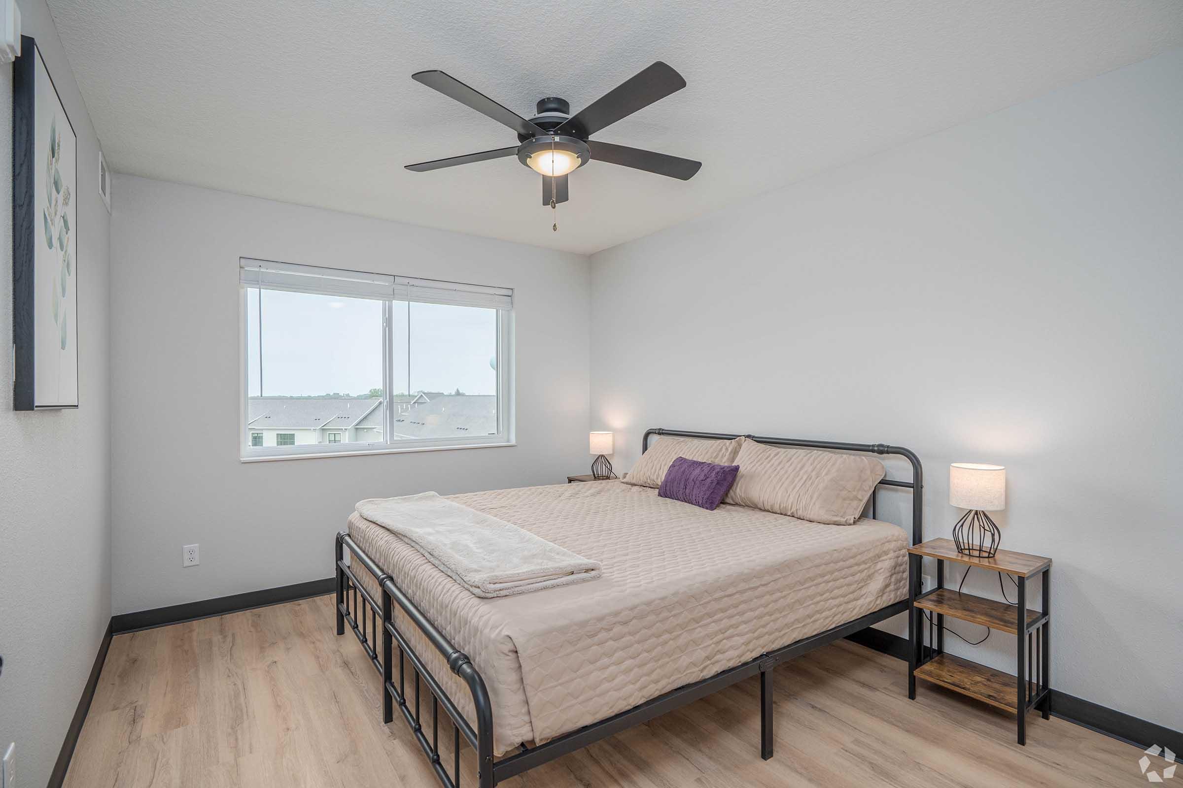 A well-lit bedroom featuring a queen-sized bed with beige bedding, a small purple pillow, and a white throw. Two bedside tables with lamps are beside the bed. A ceiling fan hangs above, and a large window shows an exterior view. The walls are light gray, and the flooring is wooden.