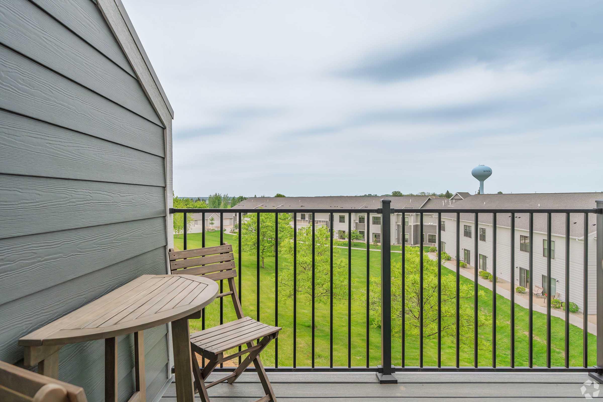 A balcony view featuring a small wooden table and two chairs. The railing overlooks a green lawn with trees and several apartment buildings in the background. A water tower is visible in the distance under a cloudy sky.