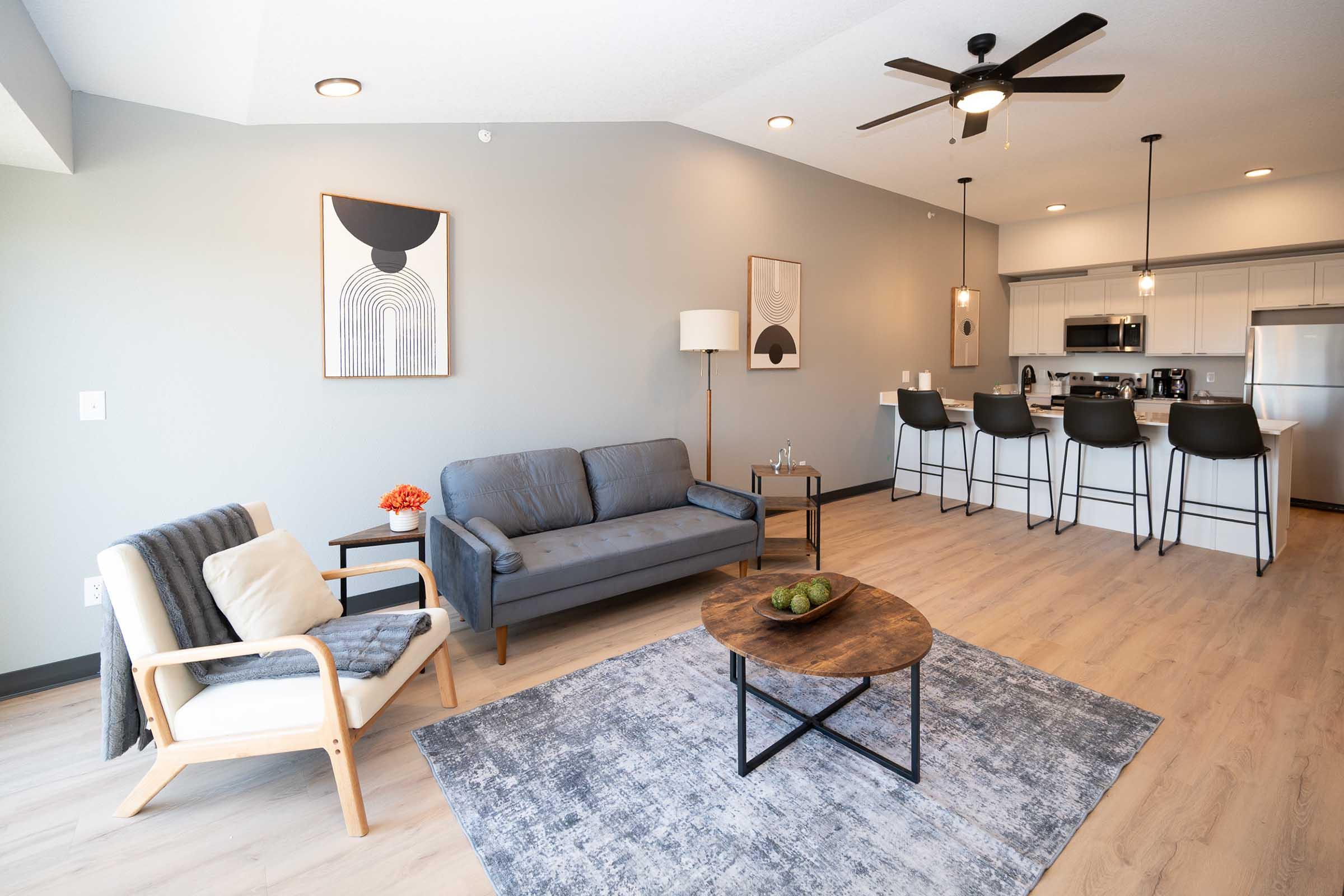 A modern living space featuring a gray sofa and an armchair with a blanket, a round coffee table, and decorative items. The room has light-colored walls, wooden flooring, and a kitchen area with bar stools. Abstract artwork is displayed on the walls, and there is soft lighting from ceiling fixtures.