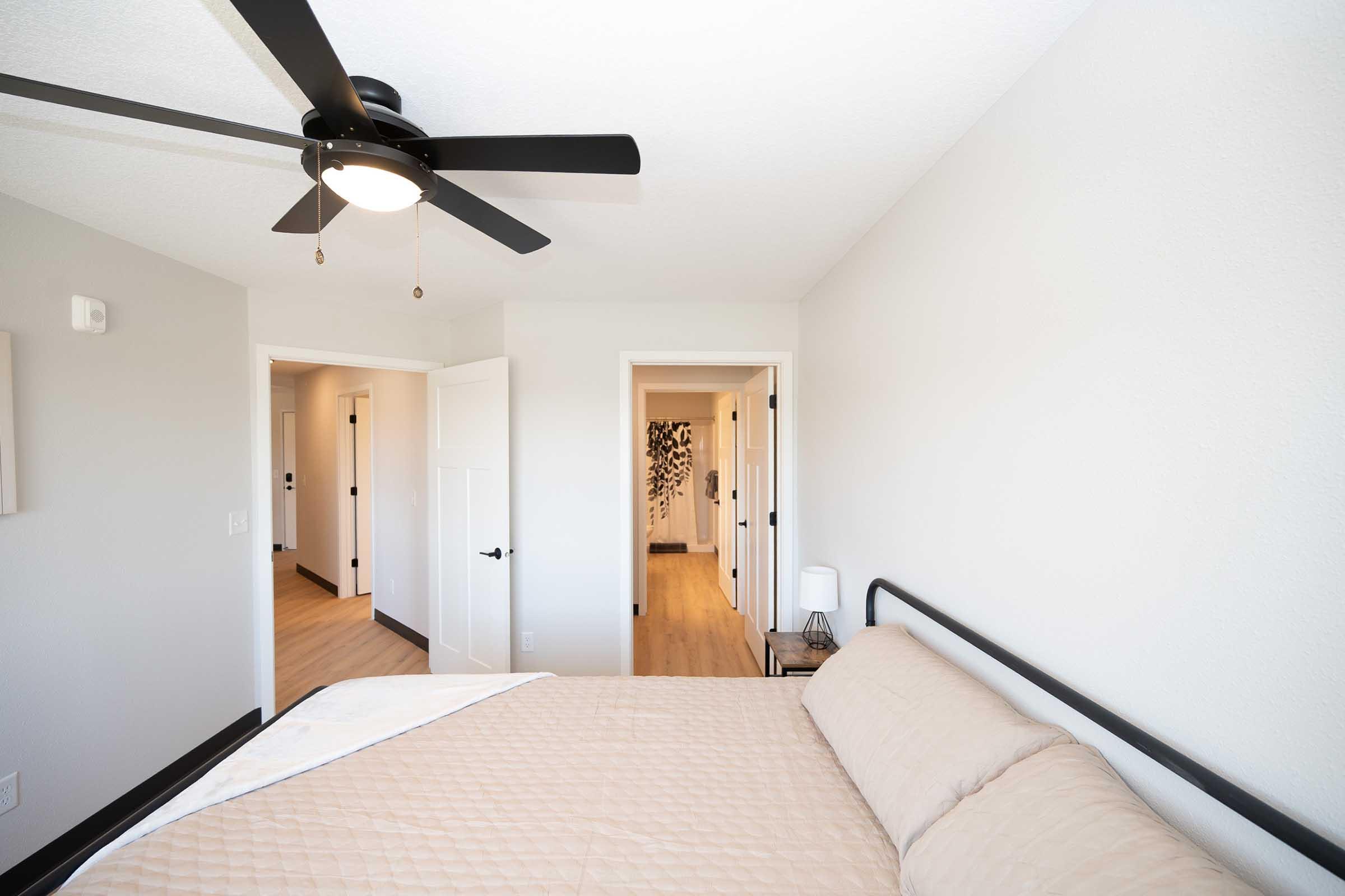 A well-lit bedroom featuring a simple metal bed with a beige comforter. The room has a ceiling fan, light-colored walls, and a modern aesthetic. Two doors lead to other areas of the home, with a glimpse of a hallway and a decorative element visible in the background.