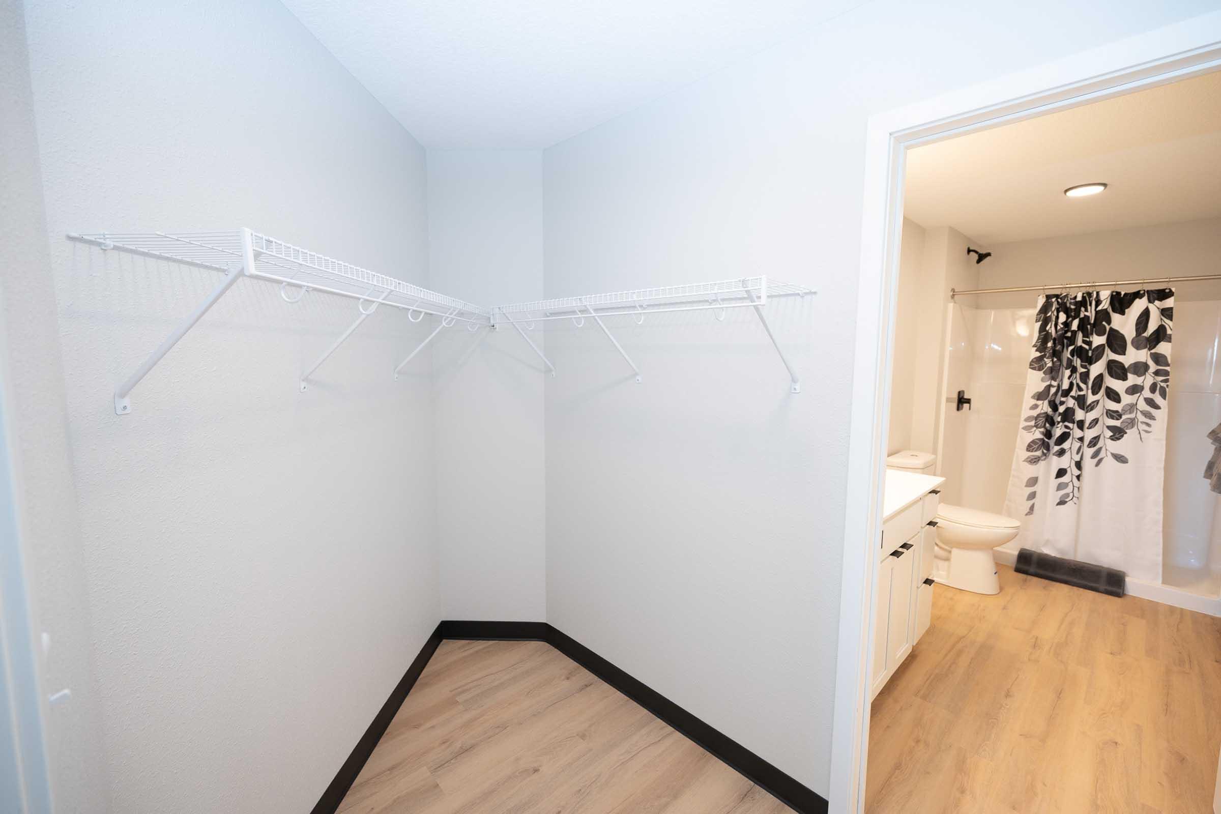 A clean and empty room with light gray walls and a wooden floor. Two wire shelving units are mounted on the wall, and a doorway leads to a bathroom featuring a shower curtain with black and white floral patterns. The bathroom includes a toilet and a sink.