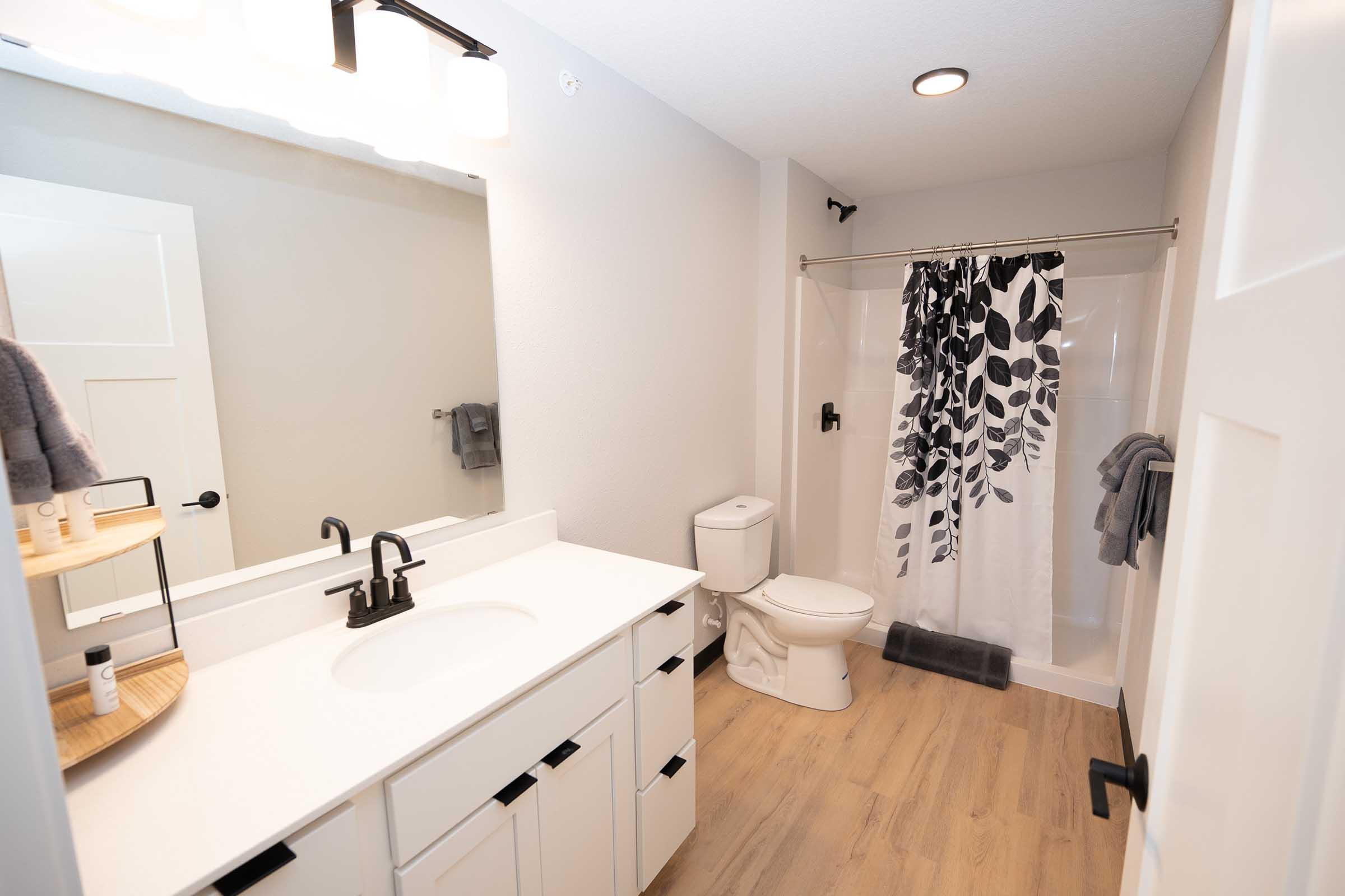 Modern bathroom featuring a white vanity with a dark faucet, a large mirror, and a shower with a black and white leaf-patterned curtain. The space has light wood flooring, a small circular shelf with toiletries, and neatly arranged towels. Soft lighting creates a bright and inviting atmosphere.