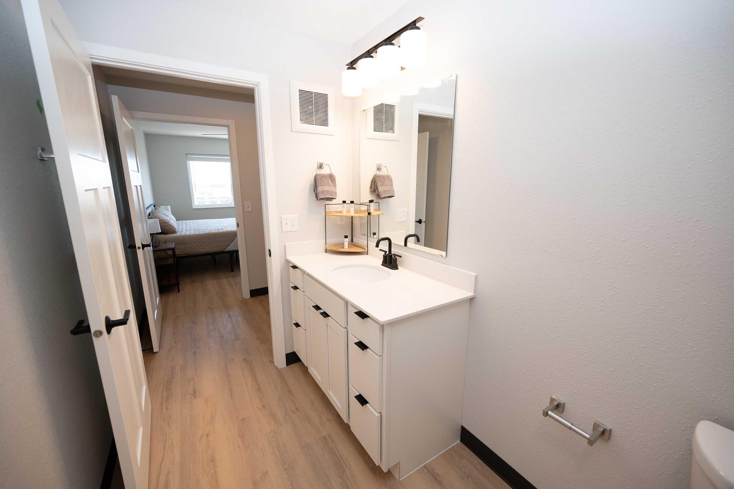 A modern bathroom featuring a white vanity with multiple drawers, a black faucet, and a large mirror. The walls are light gray, and there's wooden flooring. In the background, a doorway leads to a bedroom with natural light coming through the window. Two towels hang on the vanity.