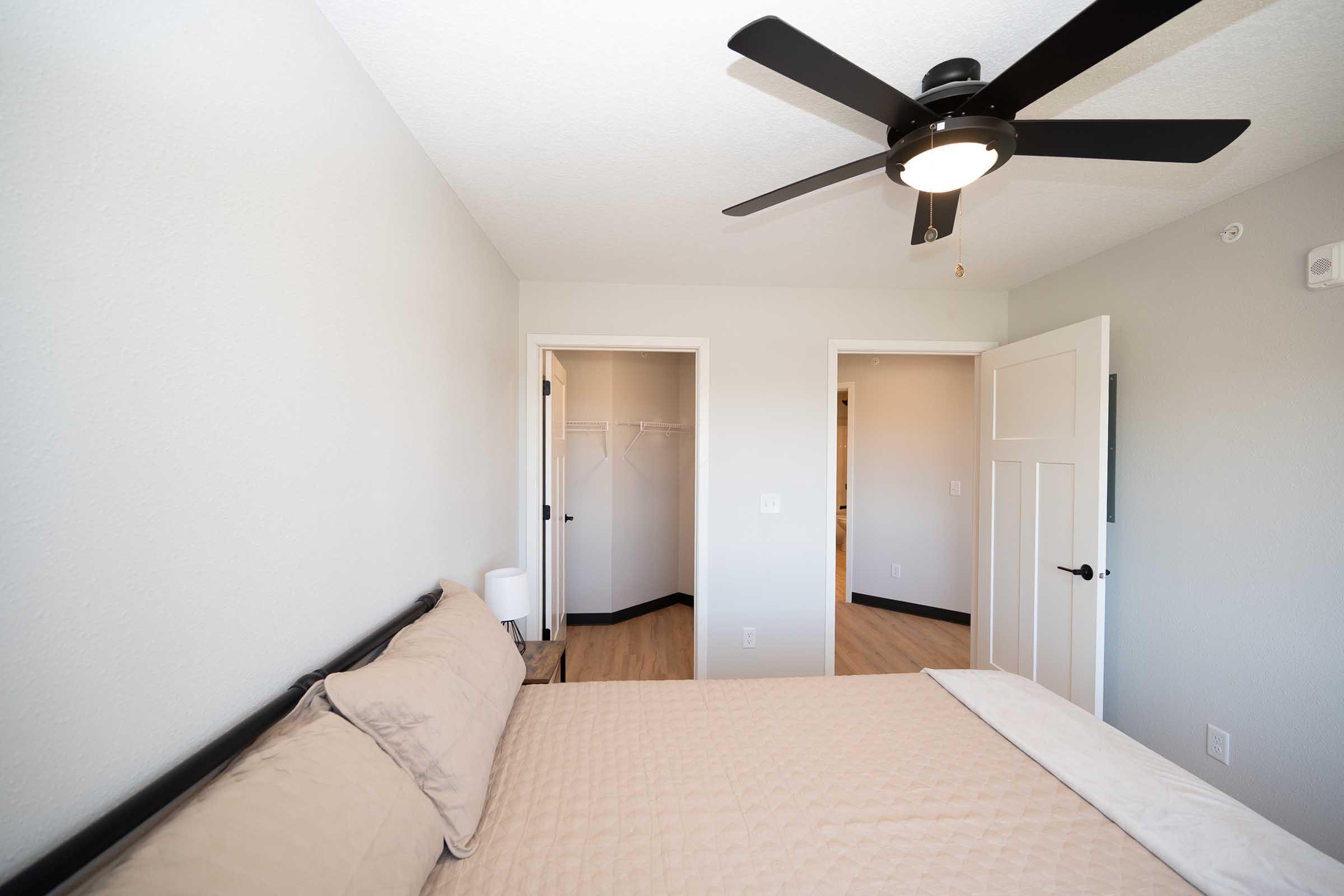 A modern bedroom featuring a light-colored wall, a ceiling fan, and a cozy bed with beige bedding. There are two doorways leading to other rooms, and a small lamp is on the nightstand. The flooring is a light wood finish, creating a bright and airy atmosphere.
