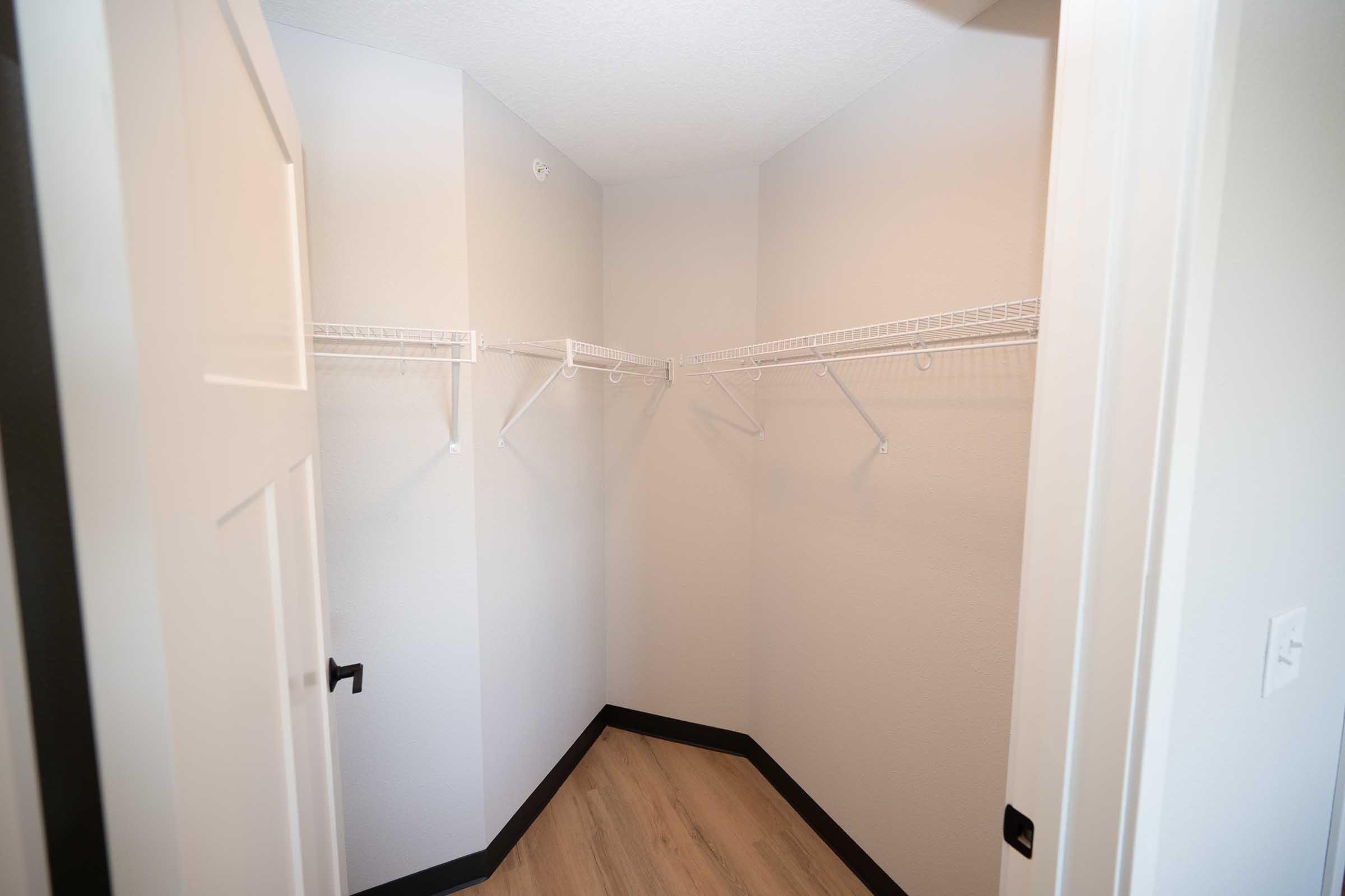 A corner closet with light gray walls and a smooth floor. The space features two wire shelving units mounted on the walls, providing storage options. A closed white door is visible on one side, and the overall lighting is bright and neutral, creating an open feel.