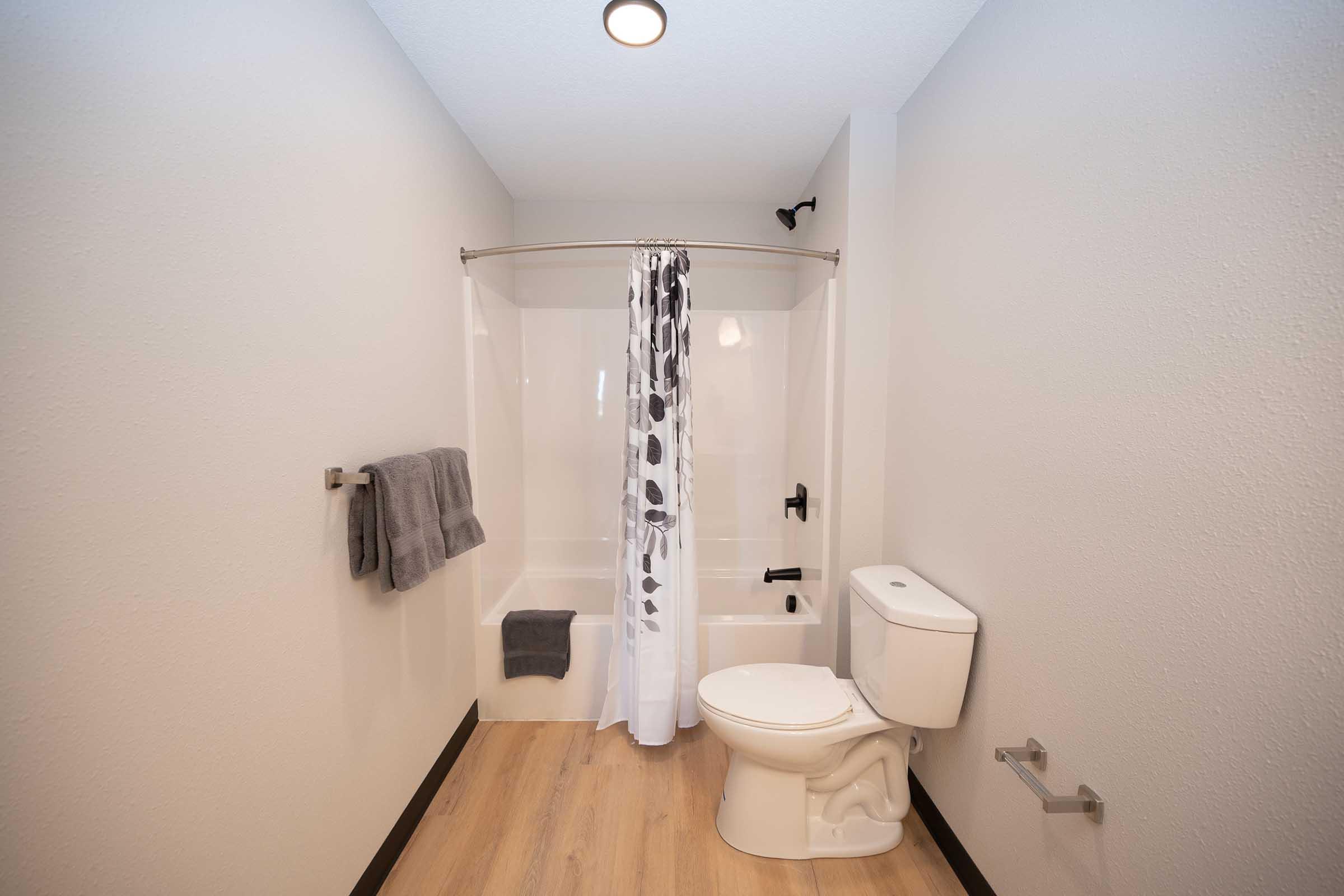 A modern bathroom featuring a white bathtub with a shower curtain, a toilet, and towels hanging on the wall. The floor has light-colored wood, and the walls are painted a neutral shade, creating a clean and minimalist aesthetic.