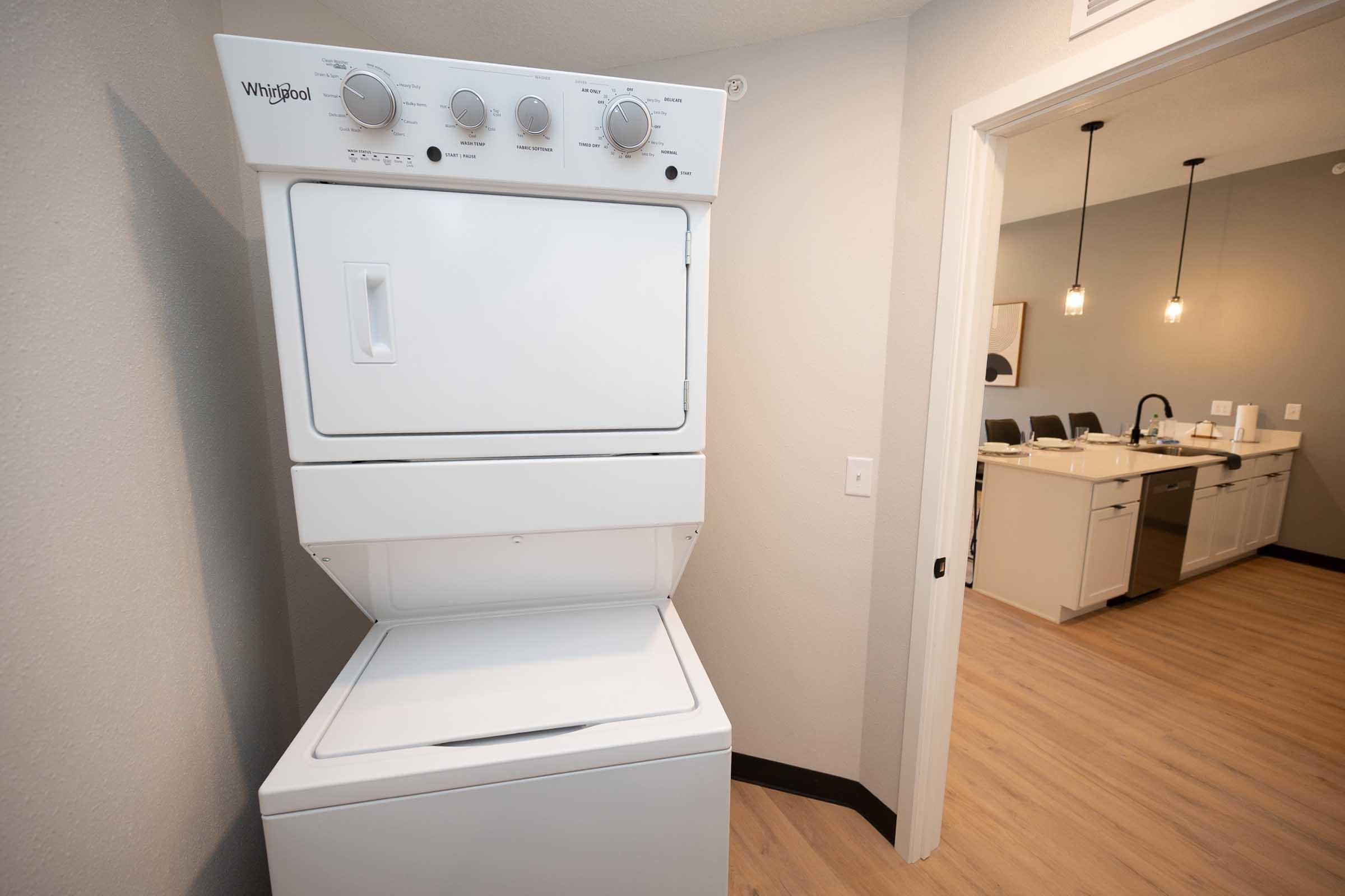 A stackable Whirlpool washer and dryer unit is installed in a light-colored room with wood-style flooring. In the background, a kitchen area is partially visible, featuring modern appliances and pendant lighting.