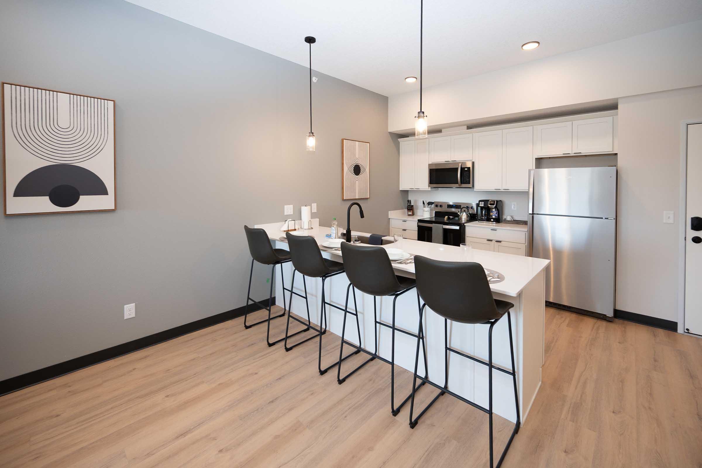 Modern kitchen with a white island countertop and four black bar stools. It features stainless steel appliances, including a refrigerator and microwave. The walls are painted a light gray, and there’s a minimalist artwork hanging above the island. Warm wood flooring complements the space.