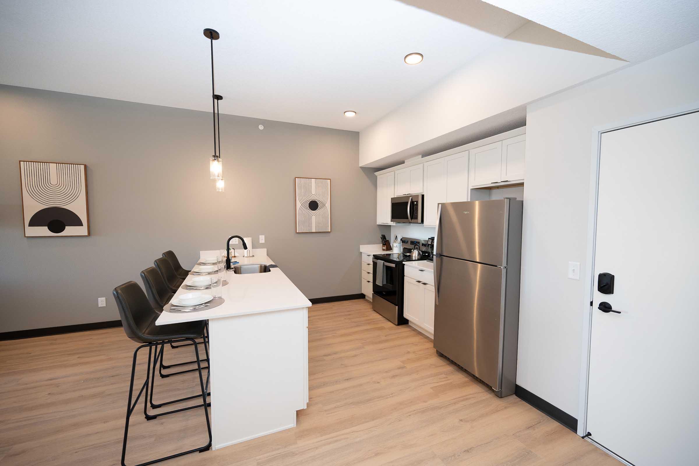 Modern kitchen featuring a spacious layout with white cabinets, stainless steel appliances, and a large island with seating for four. The walls are painted in a soft gray, complemented by contemporary artwork and stylish pendant lighting above the island, creating a bright and inviting atmosphere.