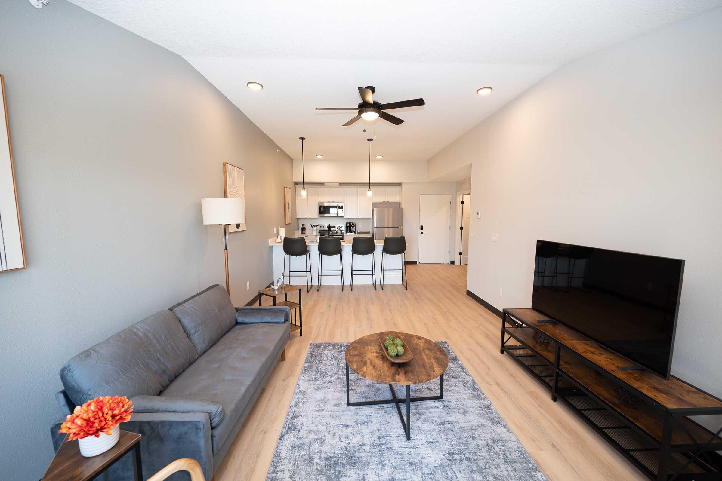 A modern living room featuring a gray sofa, a round coffee table with decorative items, and a flat-screen TV on a wooden stand. The space is bright with neutral walls, a ceiling fan, and an open kitchen area visible in the background, complete with bar stools and contemporary appliances.