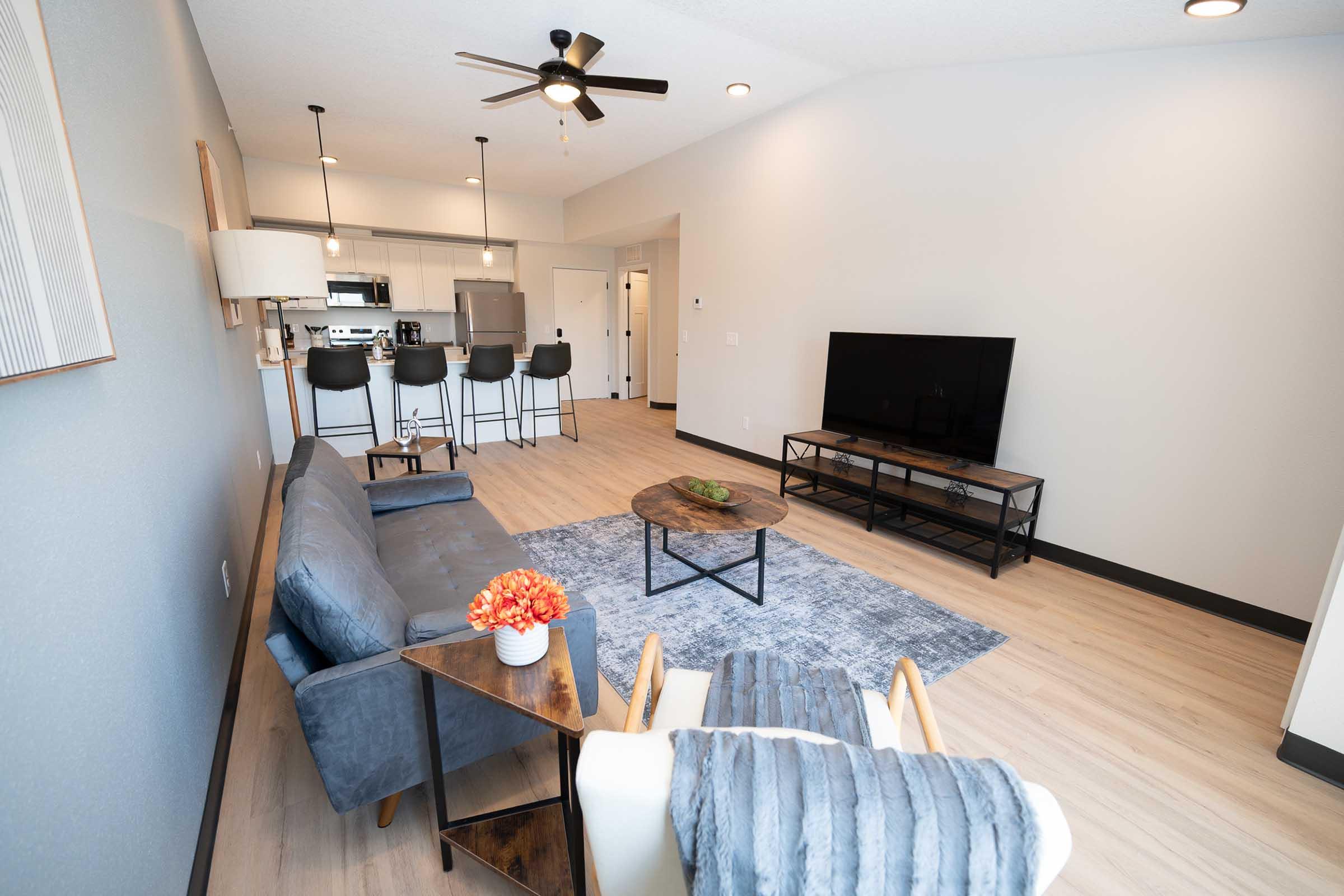 A modern living room featuring a gray sofa, a wooden coffee table, and an armchair. There's a large TV on a stand, with a rug on the floor. In the background, a kitchen area is visible with barstools and appliances. The walls are light gray, creating a bright and spacious atmosphere.