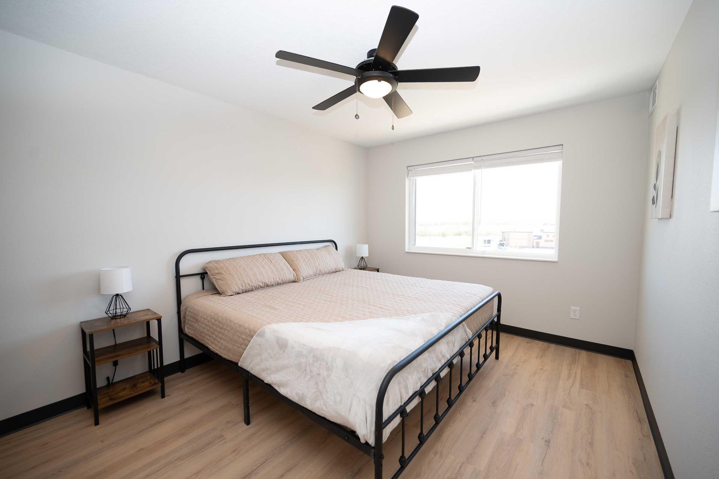 A modern bedroom featuring a large bed with a light-colored comforter, a bedside table with a lamp, and a ceiling fan. There's a window with natural light coming in, and simple decor on the walls. The flooring is light wood, creating a clean and spacious atmosphere.