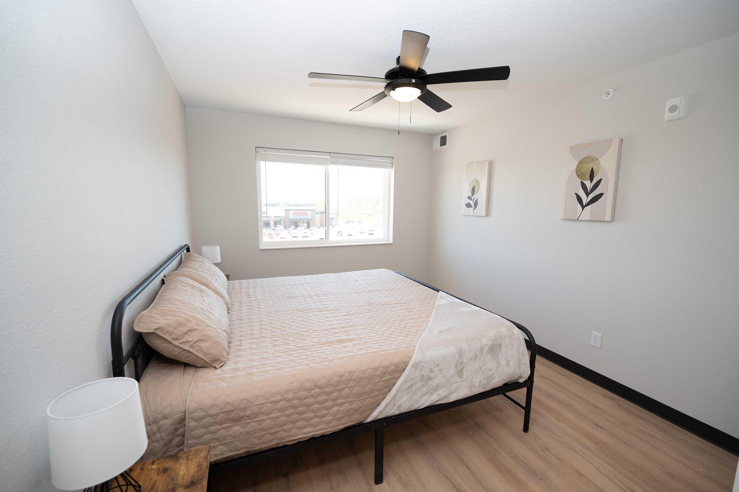 A well-lit bedroom featuring a metal-framed bed with a beige quilt, a bedside lamp, and two abstract art pieces on the wall. A ceiling fan is installed, and there's a window allowing natural light, showcasing a view of the outside. The floor is covered in light-colored laminate or wood.