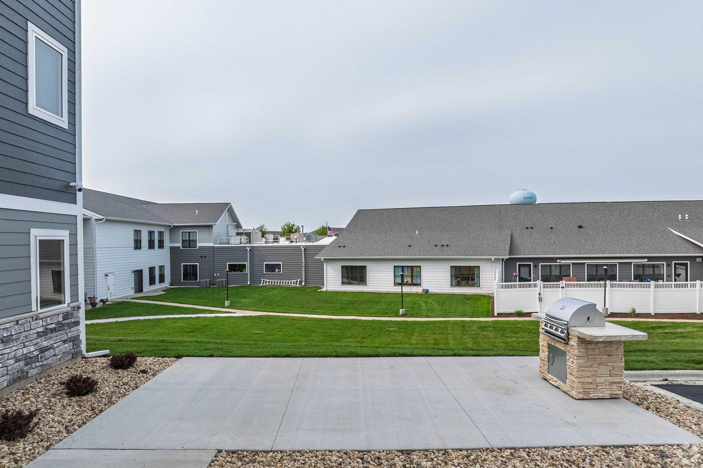 View of a residential area featuring multiple buildings with gray and white exteriors. In the foreground, a concrete pathway leads to a stone grill. The background shows a well-maintained lawn and additional neighboring buildings, under a cloudy sky.