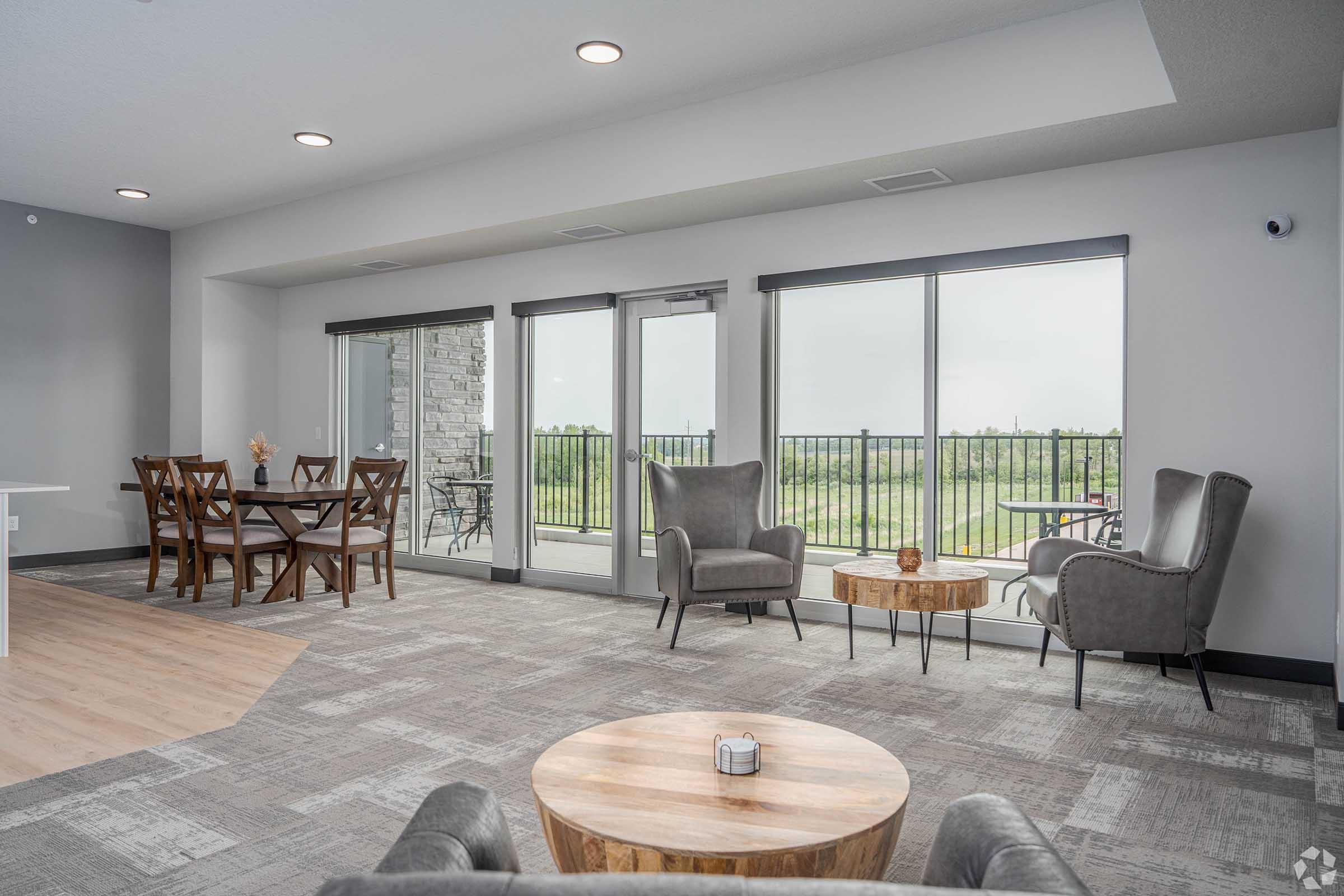 A modern, spacious interior featuring a cozy seating area with two armchairs and a small wooden coffee table. Large windows offer a view of the outdoor space, and a dining table with chairs is positioned nearby. The decor includes neutral tones and contemporary furnishings.