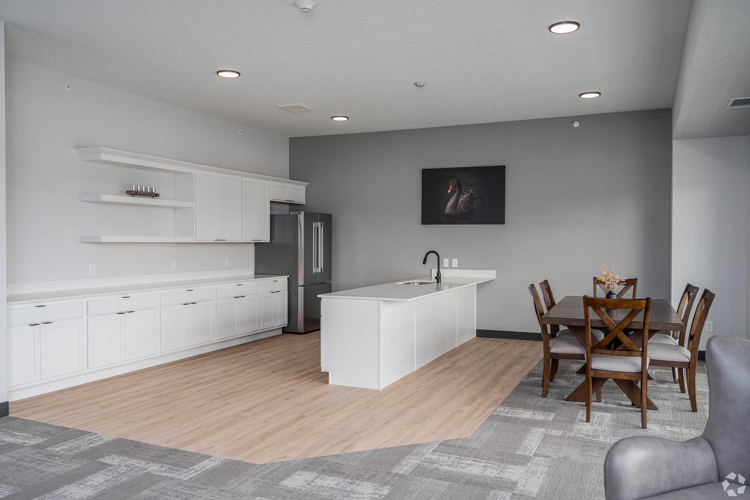 Modern kitchen and dining area in a spacious apartment featuring white cabinets, a central island with a sink, and a dining table with wooden chairs. The walls are light gray, and there is a large dark-framed artwork on one wall, with recessed lighting illuminating the space.