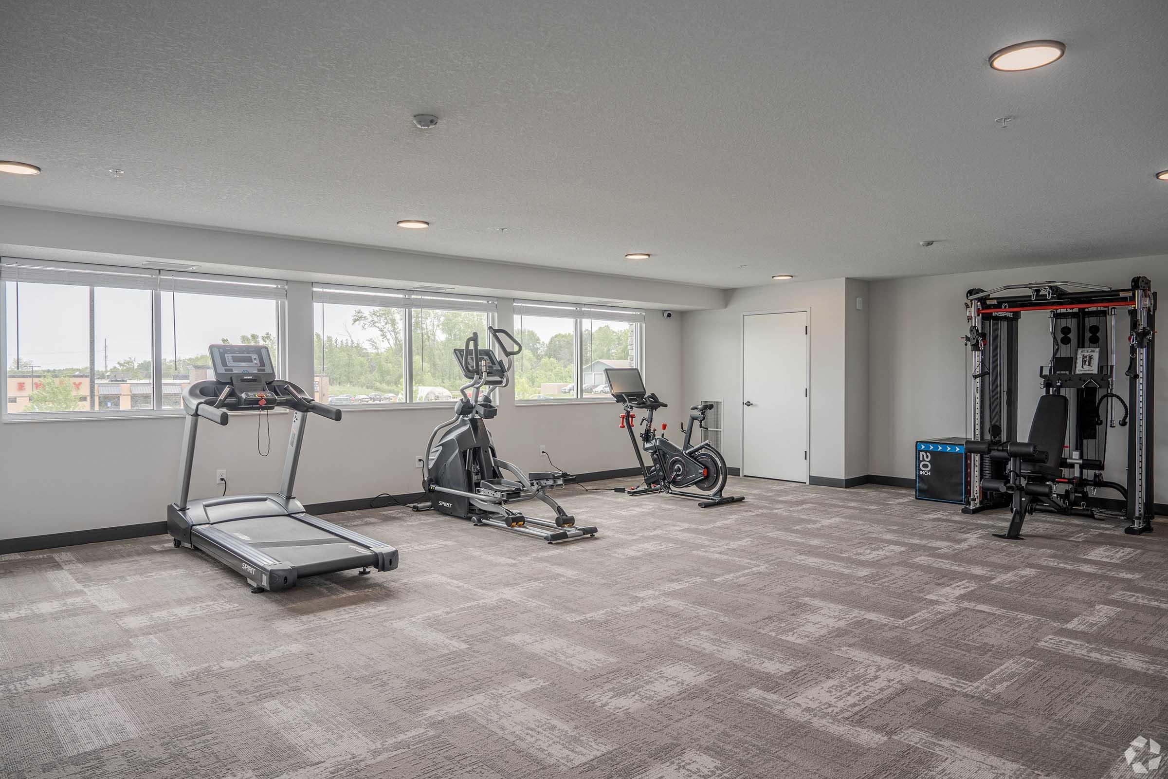A modern gym interior featuring a treadmill, elliptical machine, and stationary bike. A multi-functional weight machine is also present. The space is well-lit with large windows providing natural light, and the flooring is carpeted in a neutral tone. Walls are painted in a light color, creating an open atmosphere.