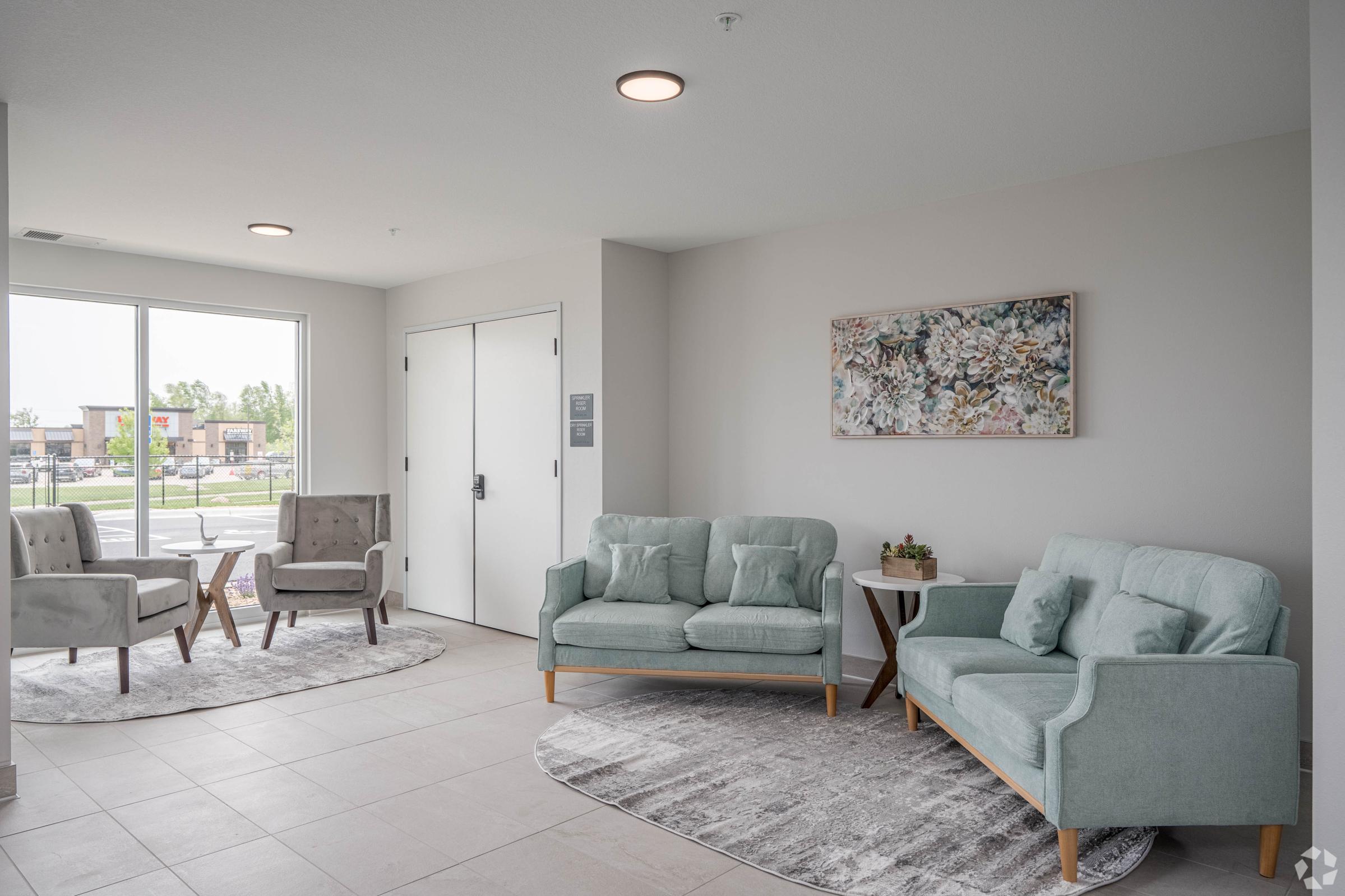 A modern waiting area featuring light-colored walls and tiled flooring. There are two light gray armchairs and a matching sofa arranged around a small side table. A round rug lies under the furniture, and a colorful artwork hangs on the wall. A large window offers a view of the outdoors.