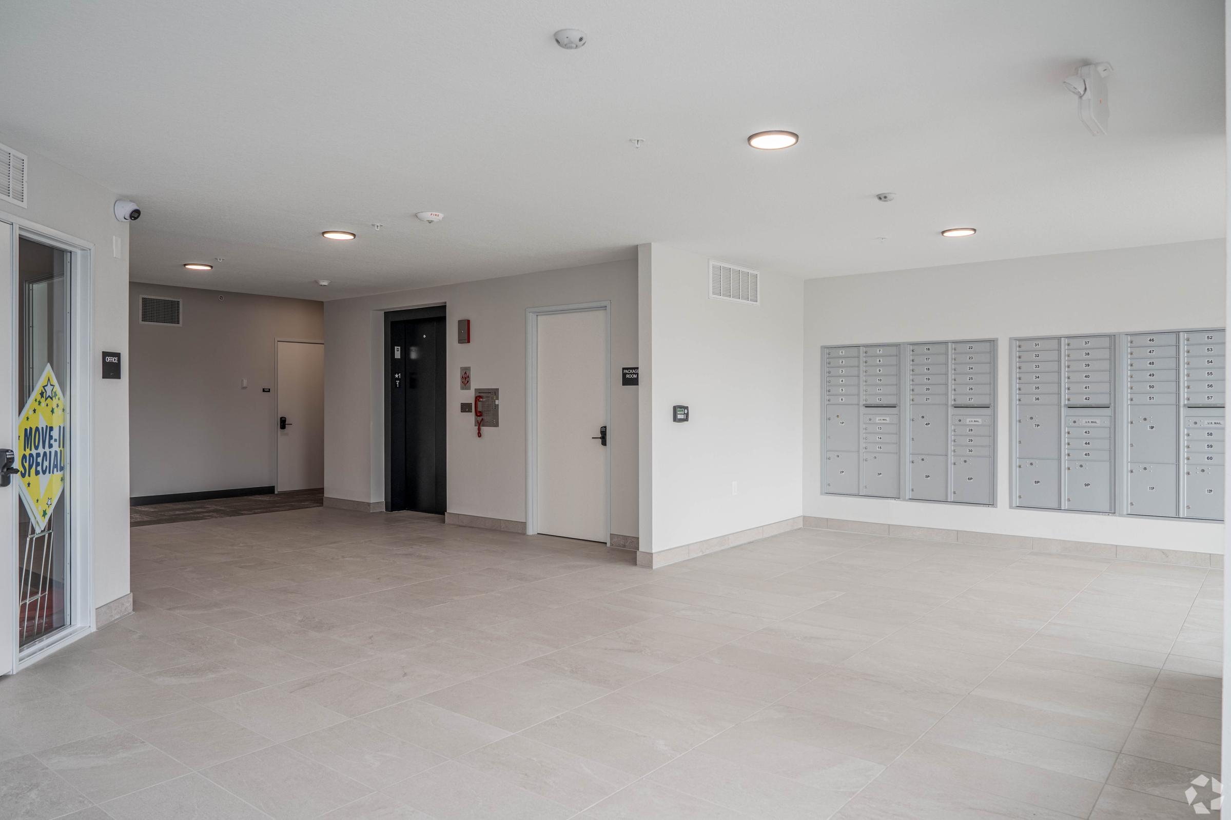 Interior of a modern building lobby featuring a light-colored tiled floor, mailboxes mounted on a wall, and an elevator in the background. A sign near the entrance indicates a special offer. The space appears bright and clean, designed for residential use.