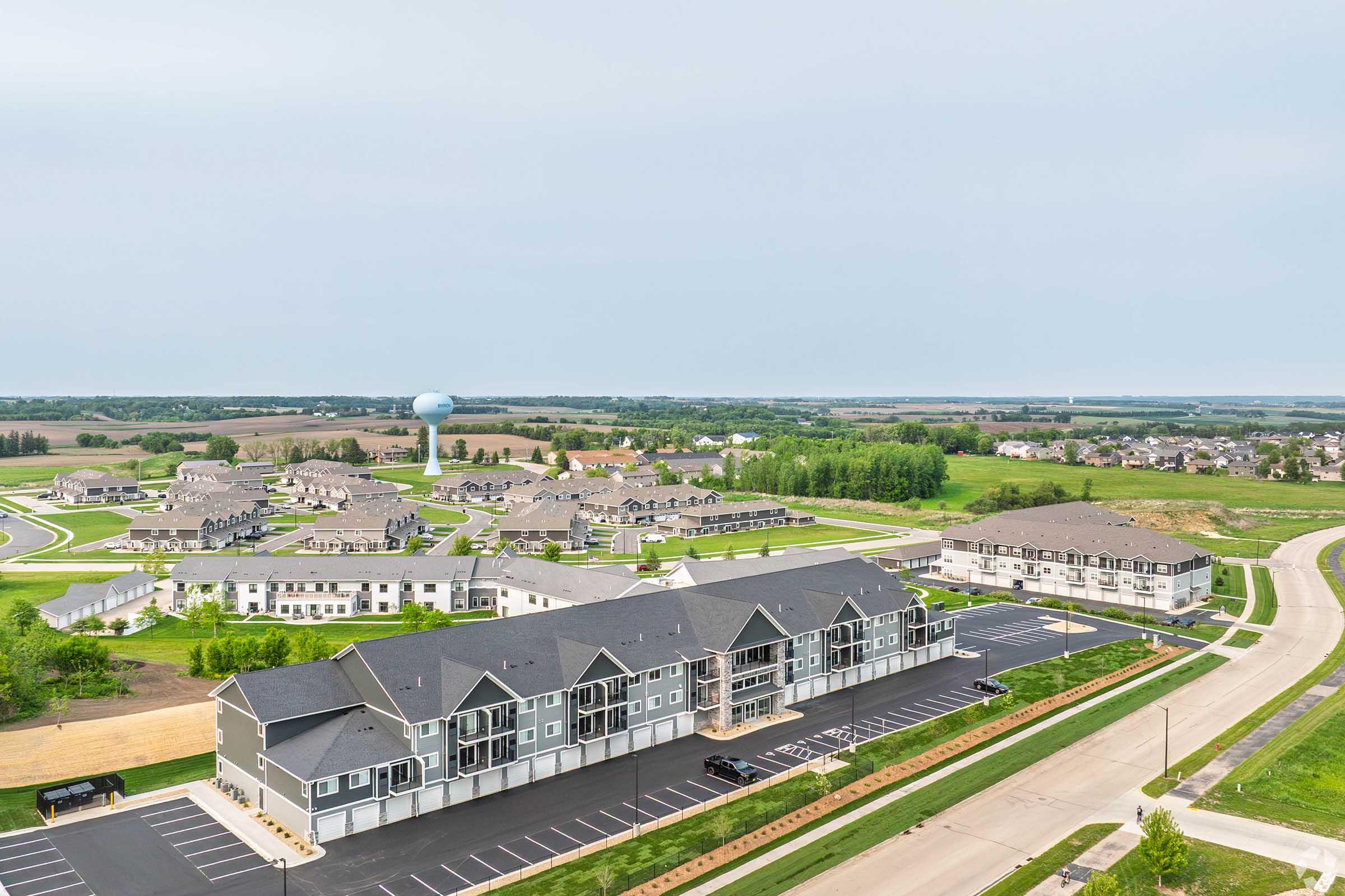Aerial view of a suburban neighborhood featuring multiple residential buildings, including townhouses and apartments, surrounded by green spaces and roads. A water tower is visible in the distance, alongside open fields and tree lines, creating a tranquil rural setting.