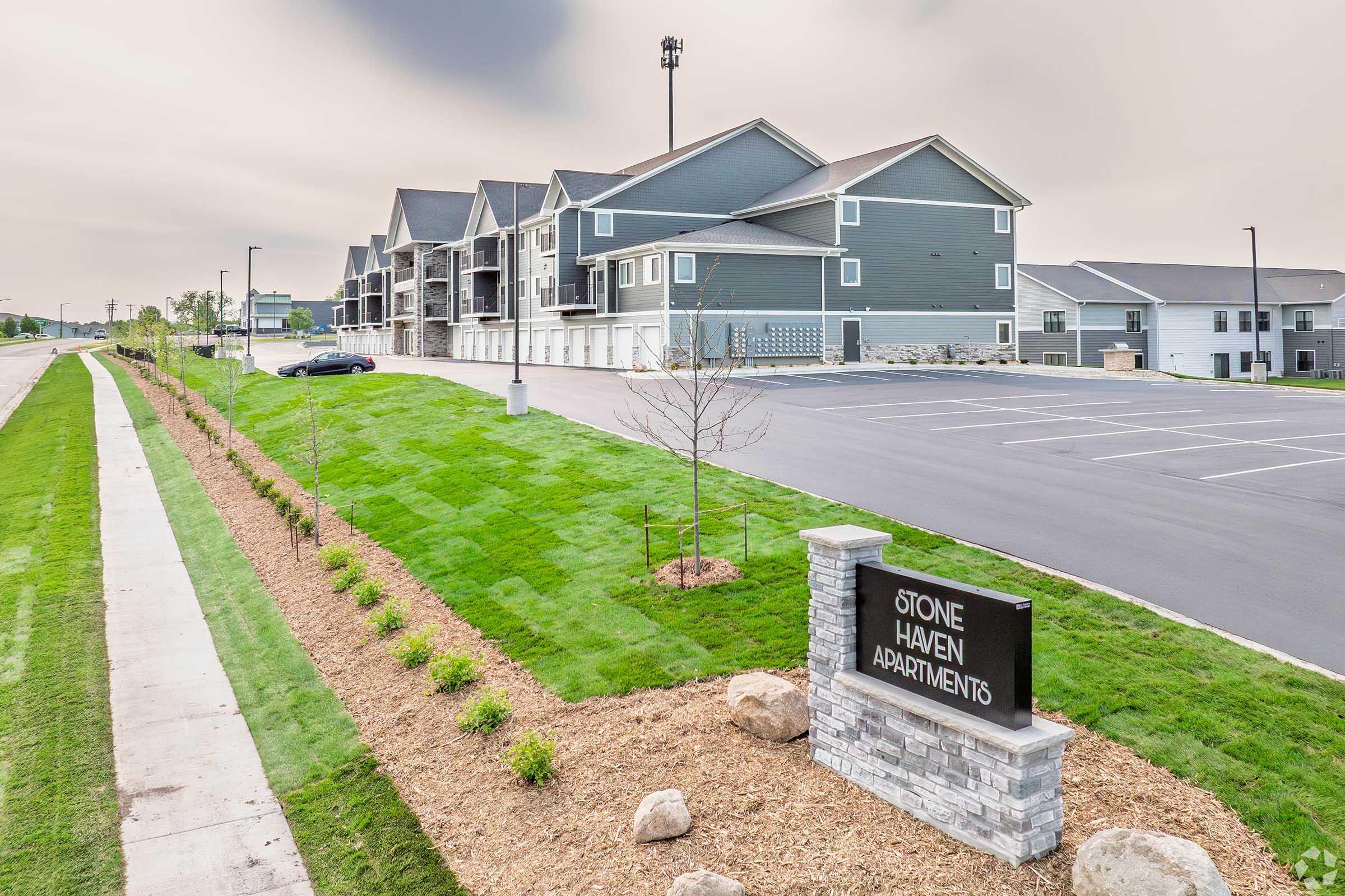 A view of Stone Haven Apartments, featuring modern multi-story buildings with gray siding. The area includes a neatly manicured lawn, a sign for the apartments, and a parking lot. The setting is suburban with a clear sky and some trees in the background.
