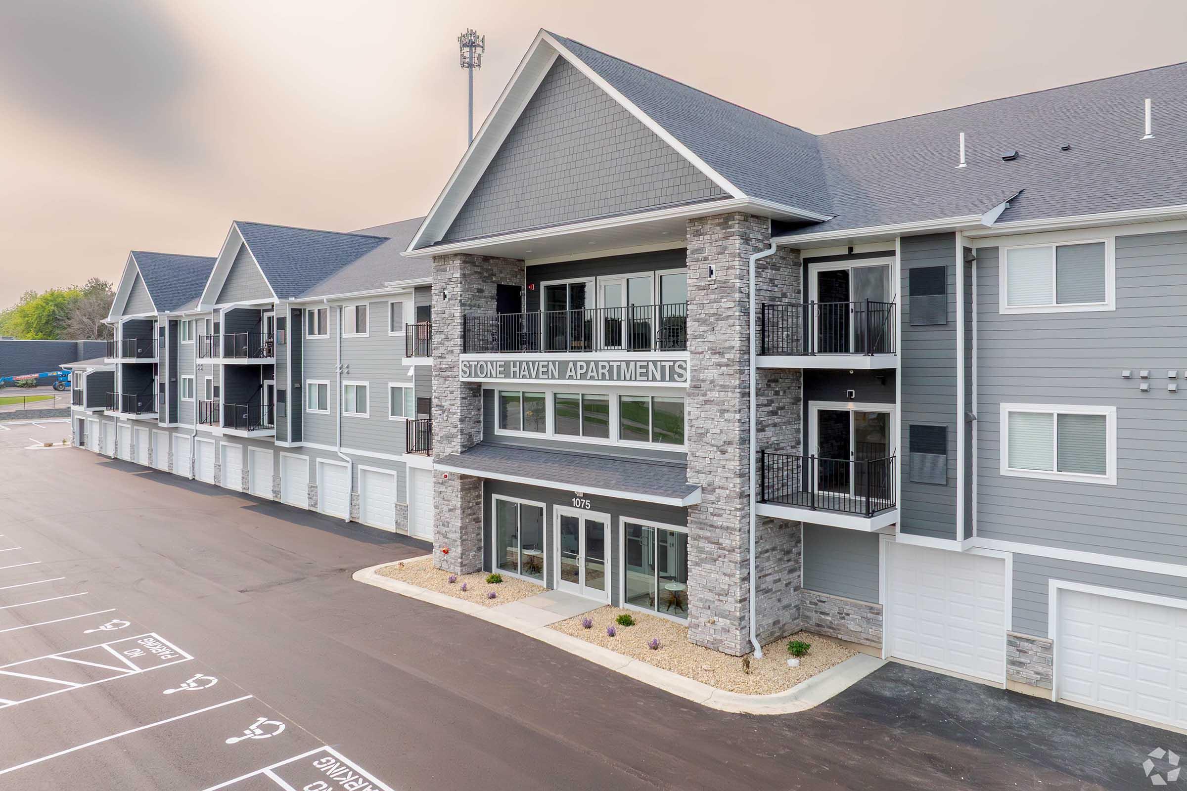 Exterior view of Stone Haven Apartments featuring a modern design with stone accents, multiple parking spaces in the foreground, and a landscaped area. The building includes balconies and large windows, set against a cloudy sky.