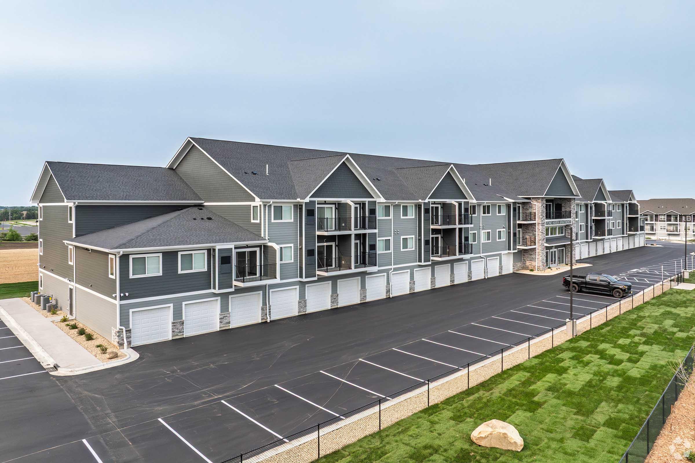 A modern residential apartment complex with multiple gray units featuring balconies, surrounded by a paved parking area. The building has stone accents and is situated near a grassy landscape and a fence. The sky is overcast, indicating a cloudy day.