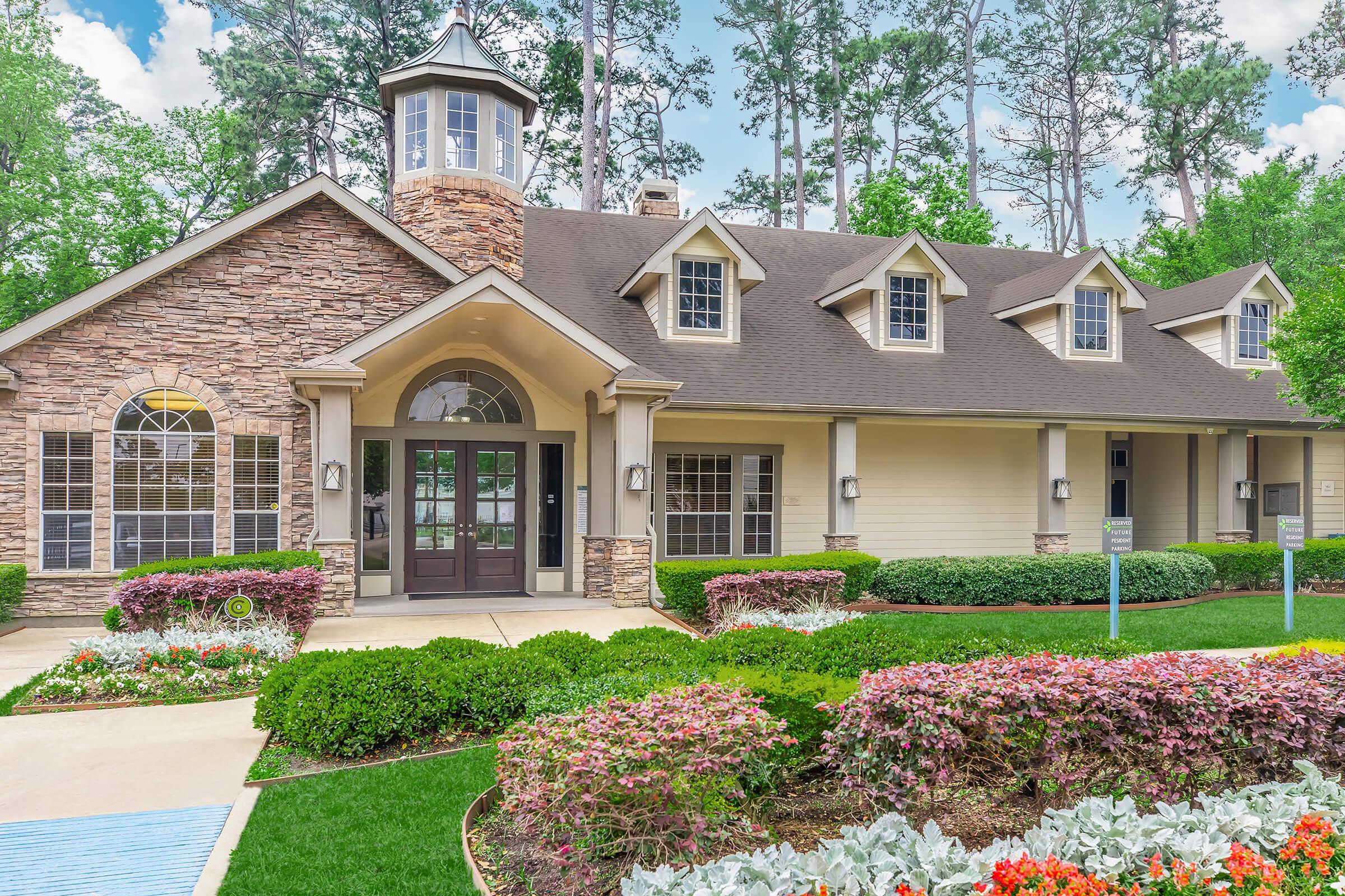 A well-maintained residential community clubhouse with a stone and siding facade, large windows, and a stylish peaked roof. Surrounding the building are lush green lawns, vibrant flower beds, and trees in the background, creating a welcoming atmosphere.