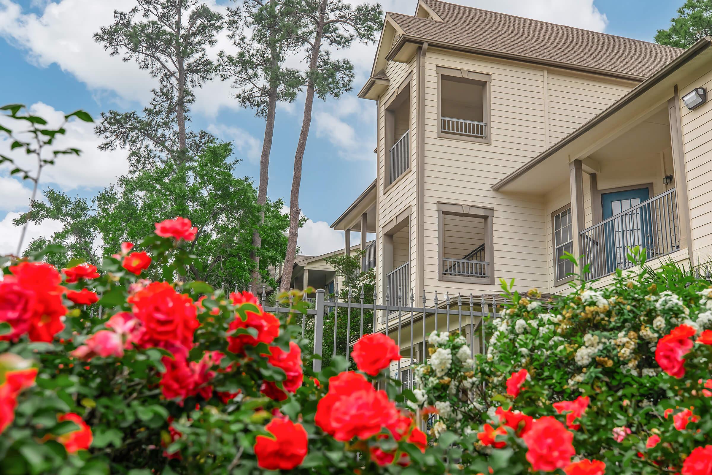A two-story light-colored house surrounded by vibrant red roses and greenery, with tall trees in the background and a clear blue sky filled with fluffy white clouds. The house features balconies and large windows, showcasing a welcoming atmosphere.