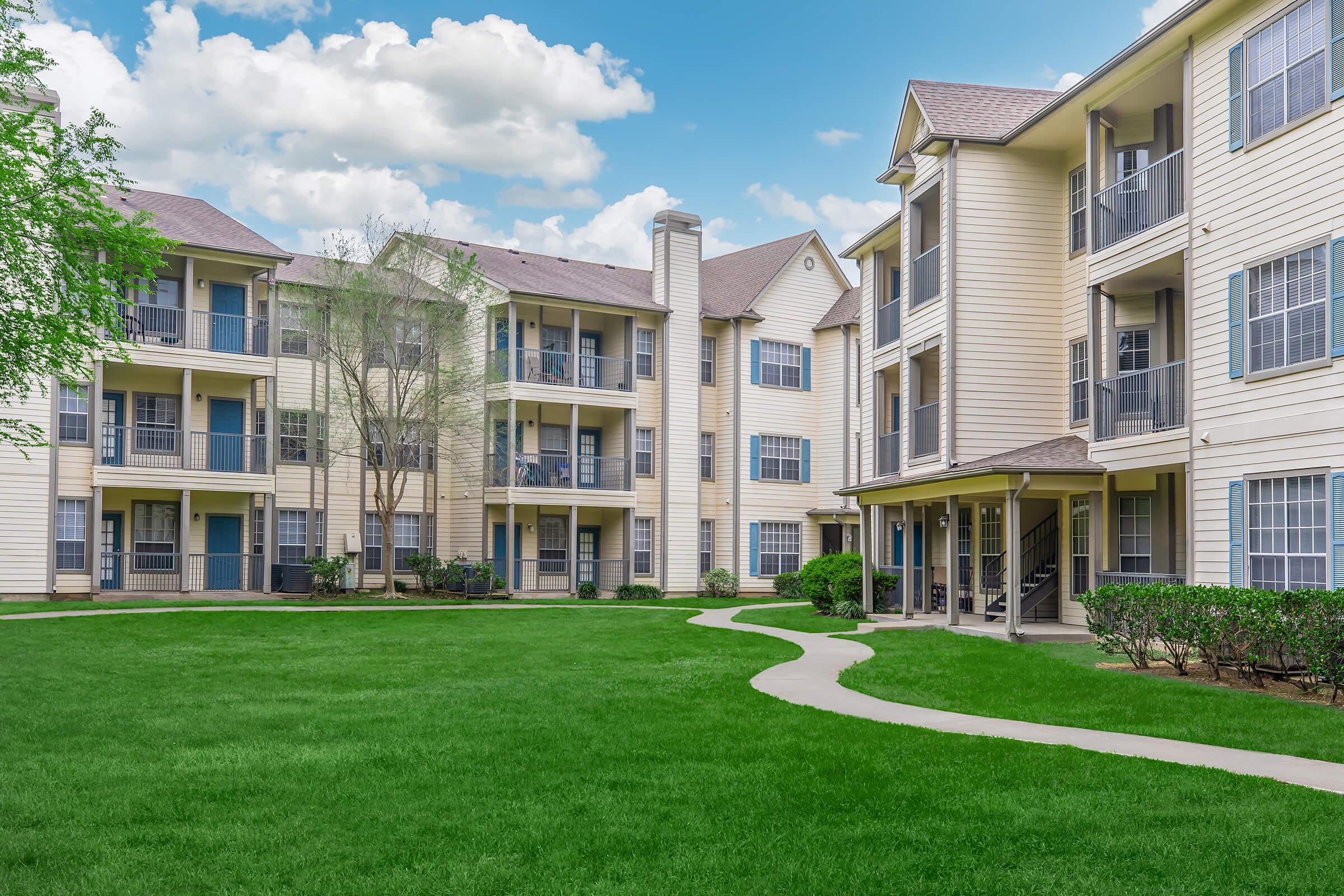 A well-maintained apartment complex with two buildings surrounding a green lawn. The architecture features multiple balconies and large windows, set against a clear blue sky with a few clouds. A winding pathway leads through the lawn, enhancing the inviting outdoor space.