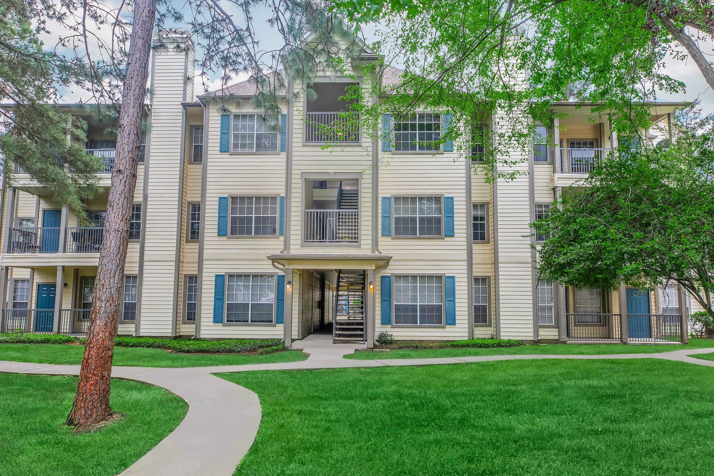 A multi-story apartment building with a cream-colored exterior, blue shutters, and balconies. The entrance is framed by trees and landscaped grass, with a curved pathway leading up to the building. The setting is bright and inviting, suggesting a pleasant residential community.