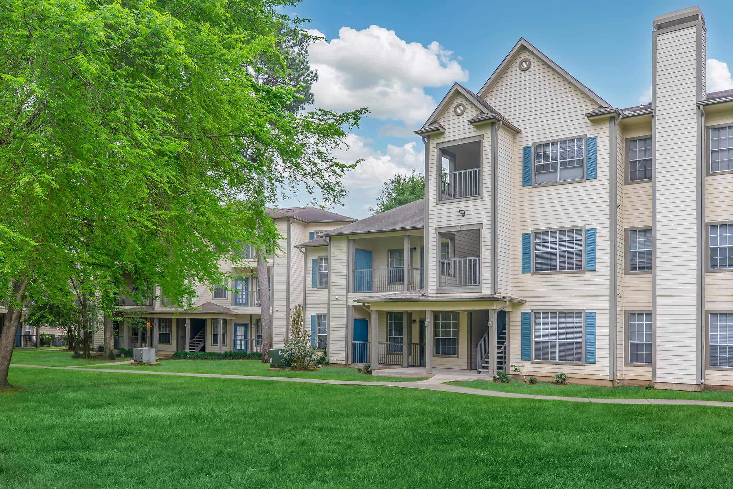 A two-story apartment building with light-colored siding, featuring multiple balconies and large windows. The surrounding area includes well-maintained green grass and trees, with a clear blue sky in the background. Steps lead to the entrance of the building, enhancing accessibility.
